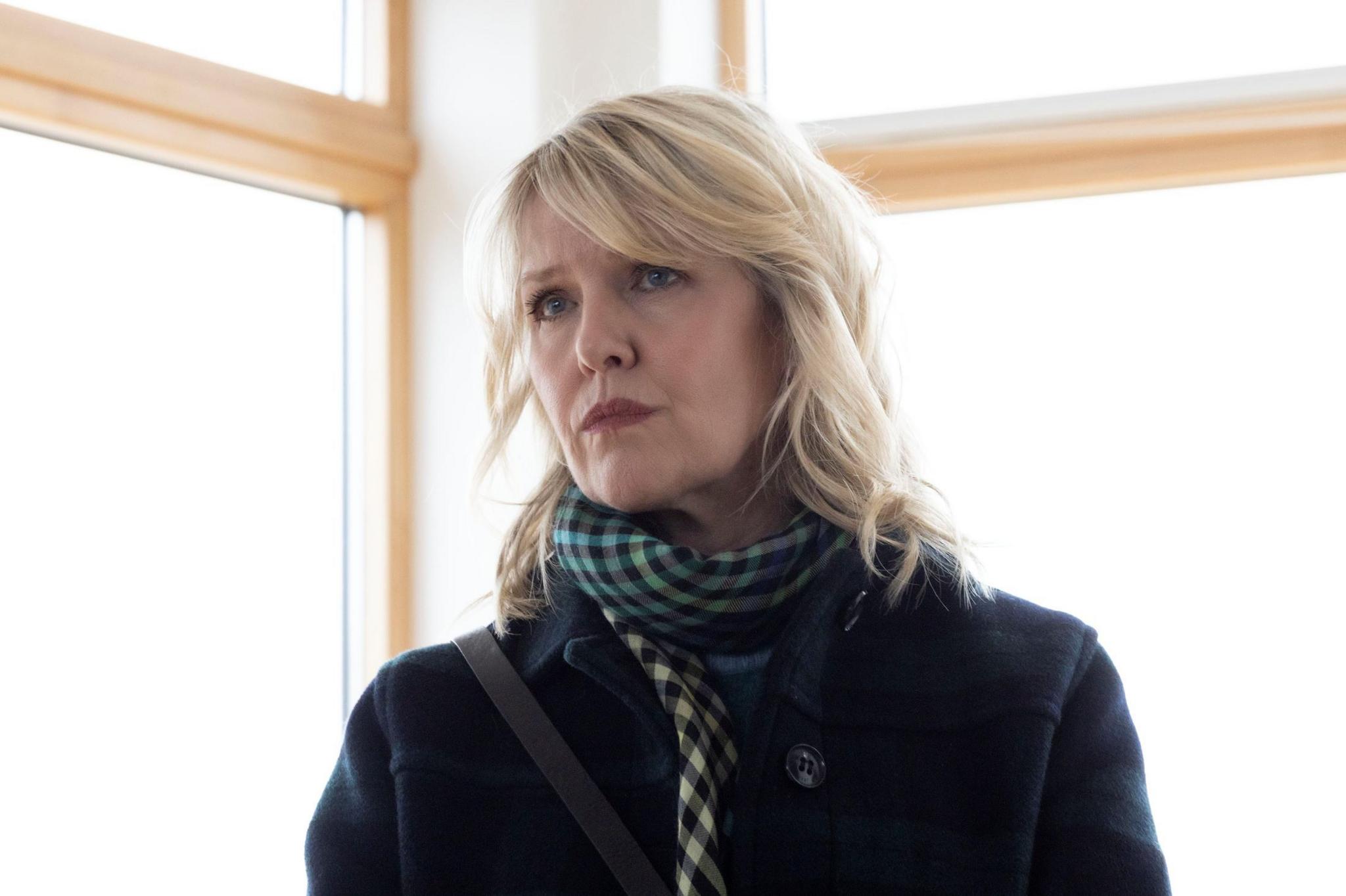 Ashley Jensen looks off to the right with concern on her face. Bright light floods in through a corner window behind her. She is wearing her dark coat and patterned scarf