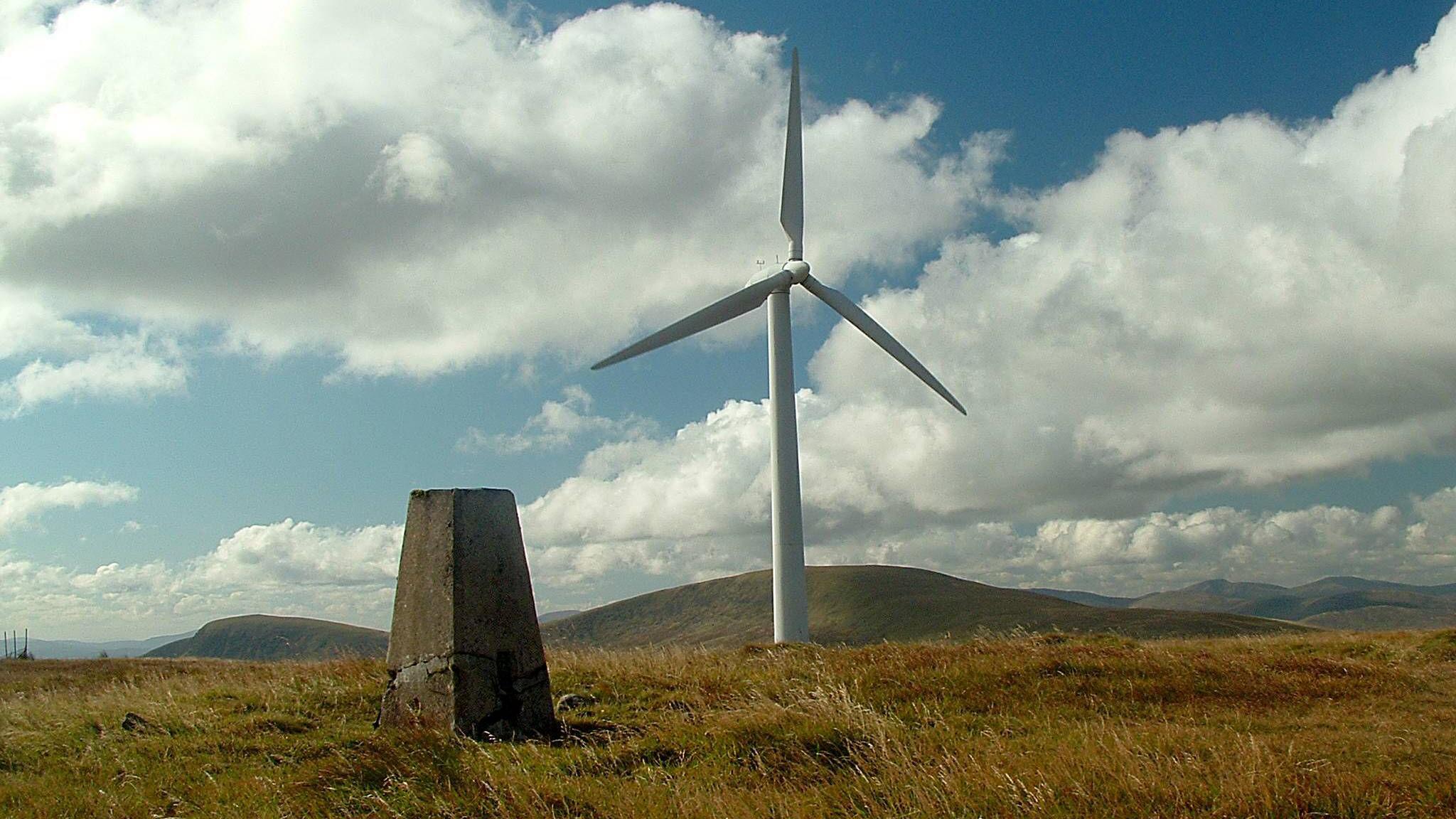 A lone wind turbine stands next to a stone marker in the Galloway hills on a cloudy day with some blue skies peeping through