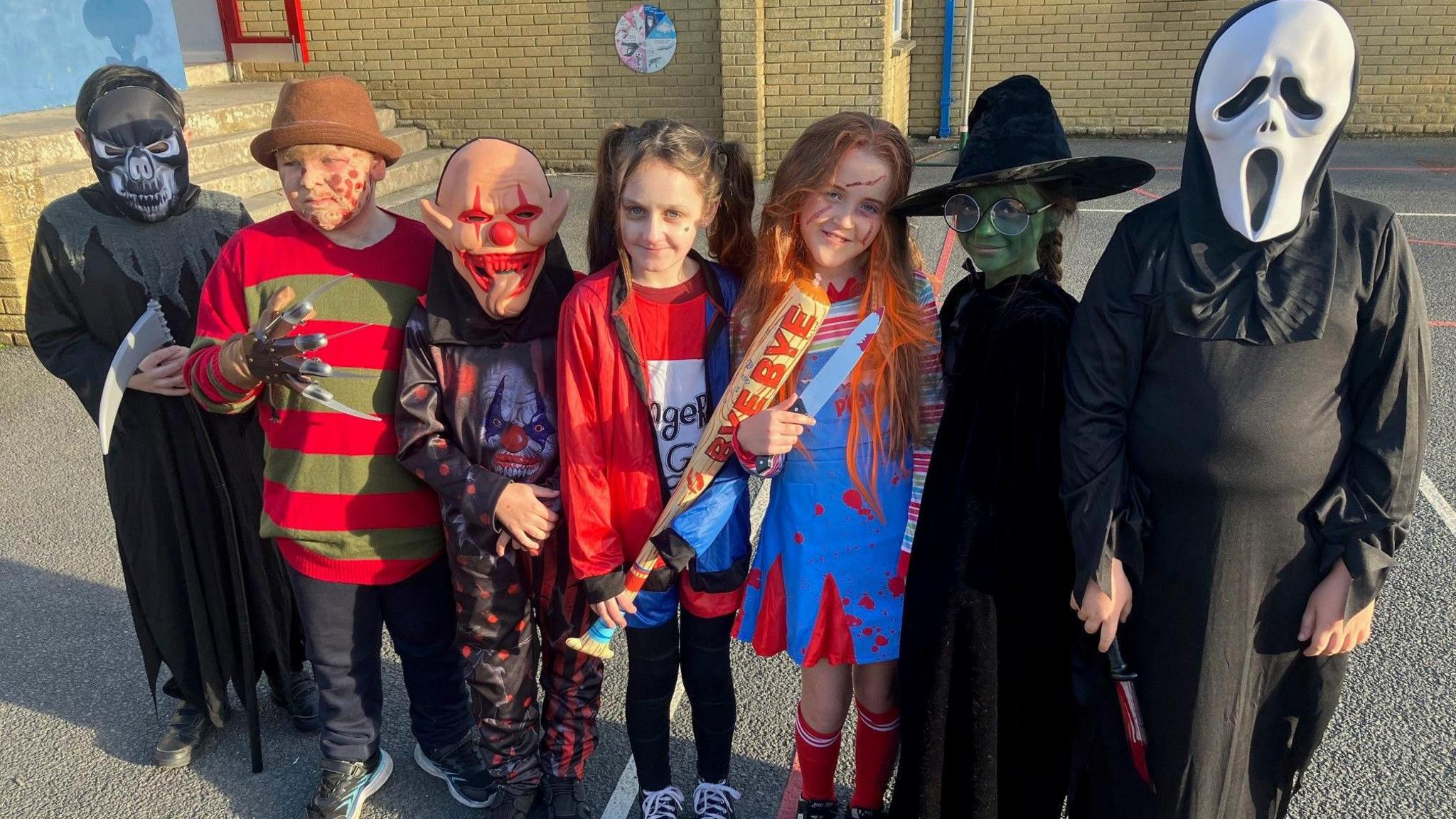 Children are dressed up in Halloween costumes. From left to right, there is a grim reaper, Freddie Kruger, a scary clown with a tongue hanging out of its face, Harley Quinn holding a baseball bat, a Chuckie with blood spattered on her dress, Elphaba from the film Wicked and Ghostface from the Scream films. They are standing is a playground with a school building behind them.