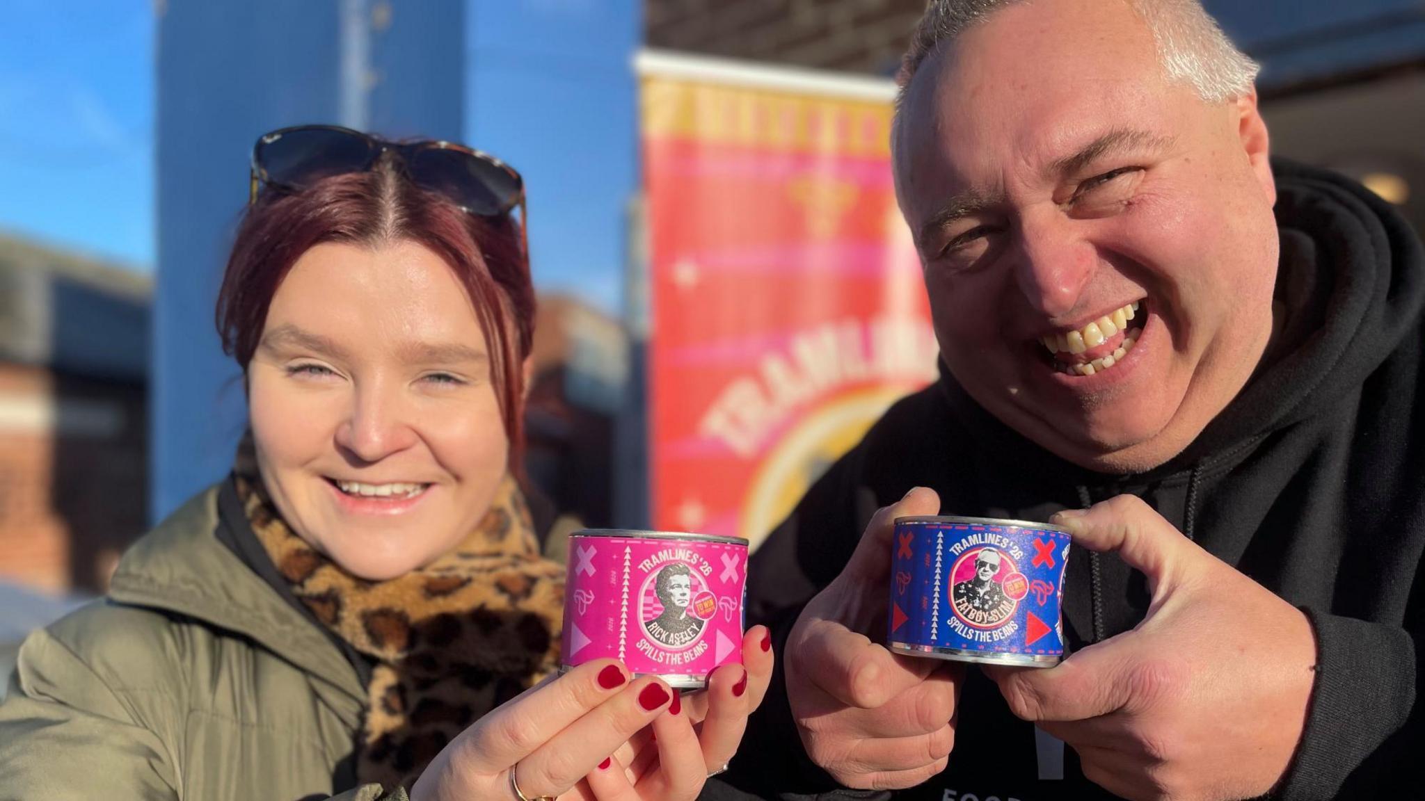 A woman with red hair wearing a thick green winter coat stands next to a jolly-looking man in a black hoodie. They are both holding up miniature cans of beans featuring pictures of Rick Astley and Fatboy Slim.