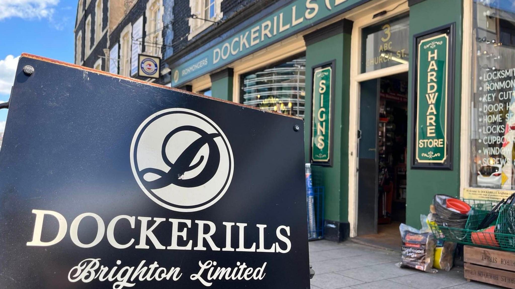 A sign for the former Dockerills store. The sign is black with white writing and there is a green building in the background. 