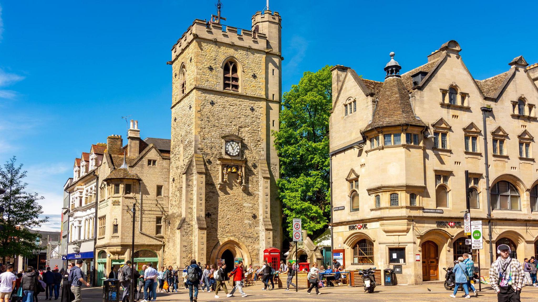 A general view of Carfax Tower at the corner of Cornmarket and Queen Street in Oxford. There are blue skies around it and people walking around the two streets below