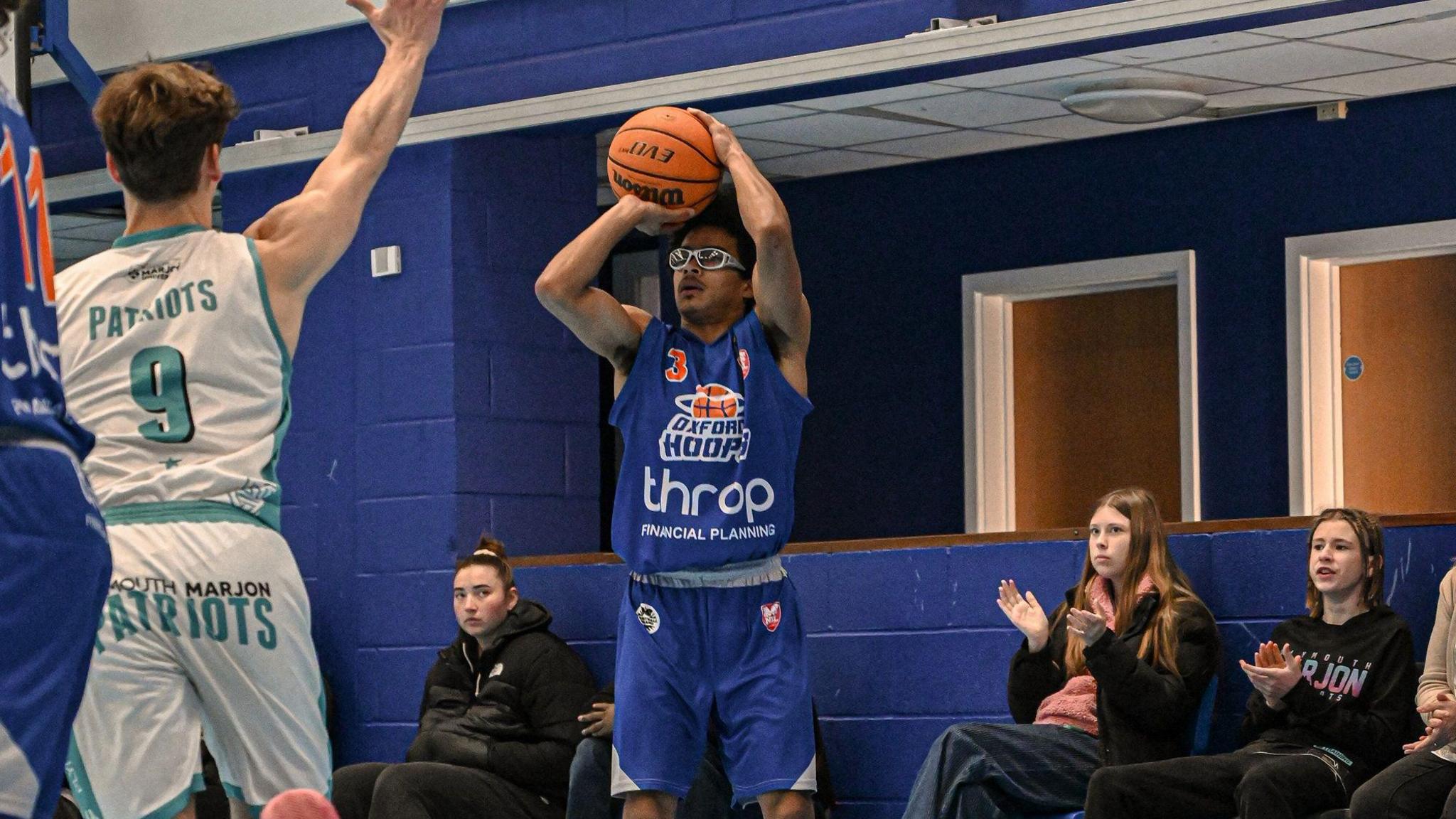 An Oxford Hoops player, wearing a blue jersey, is cocking the ball back to shoot at the hoop.