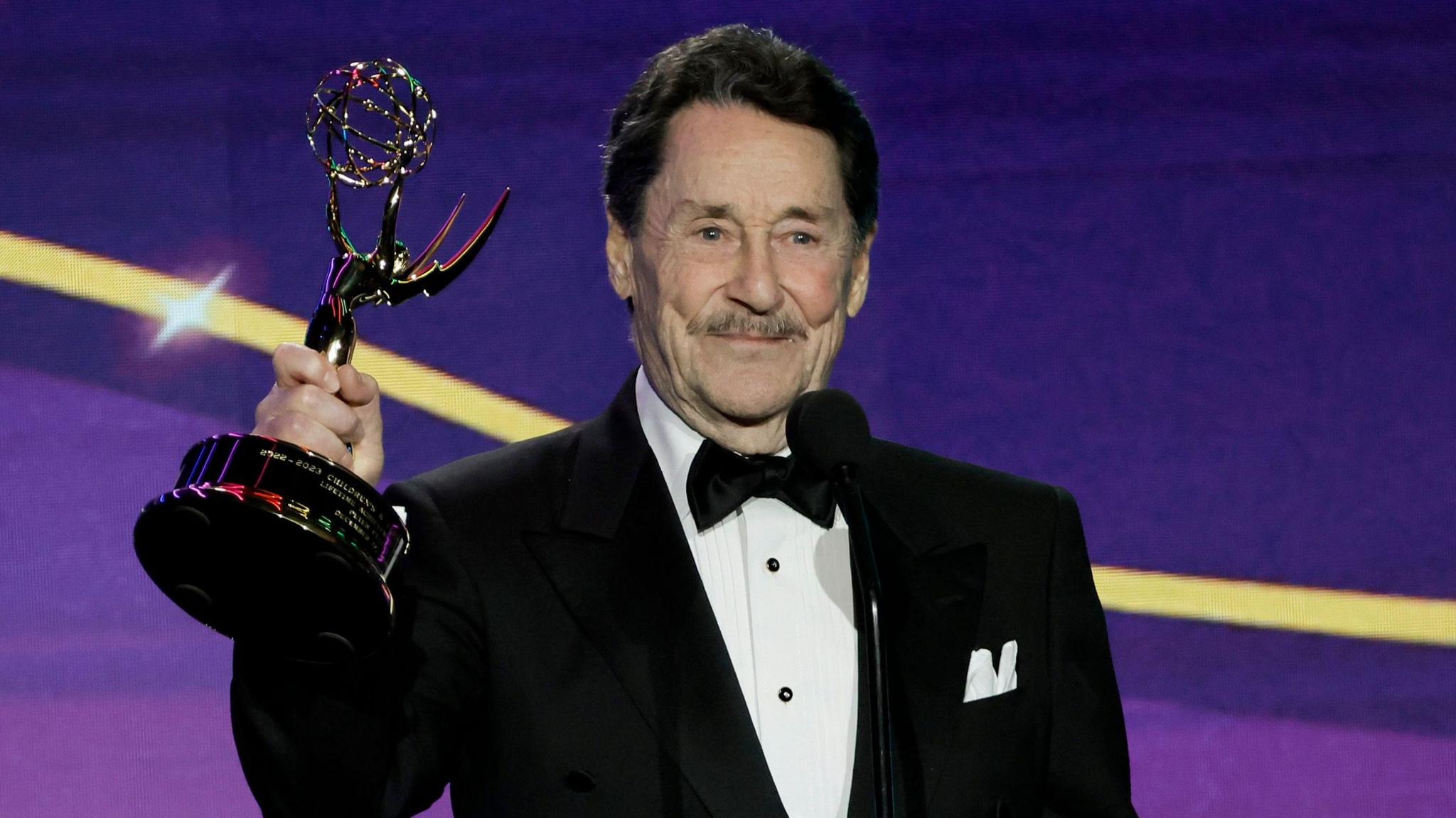 Peter Cullen in a dinner jacket holding up his Emmy Award on stage
