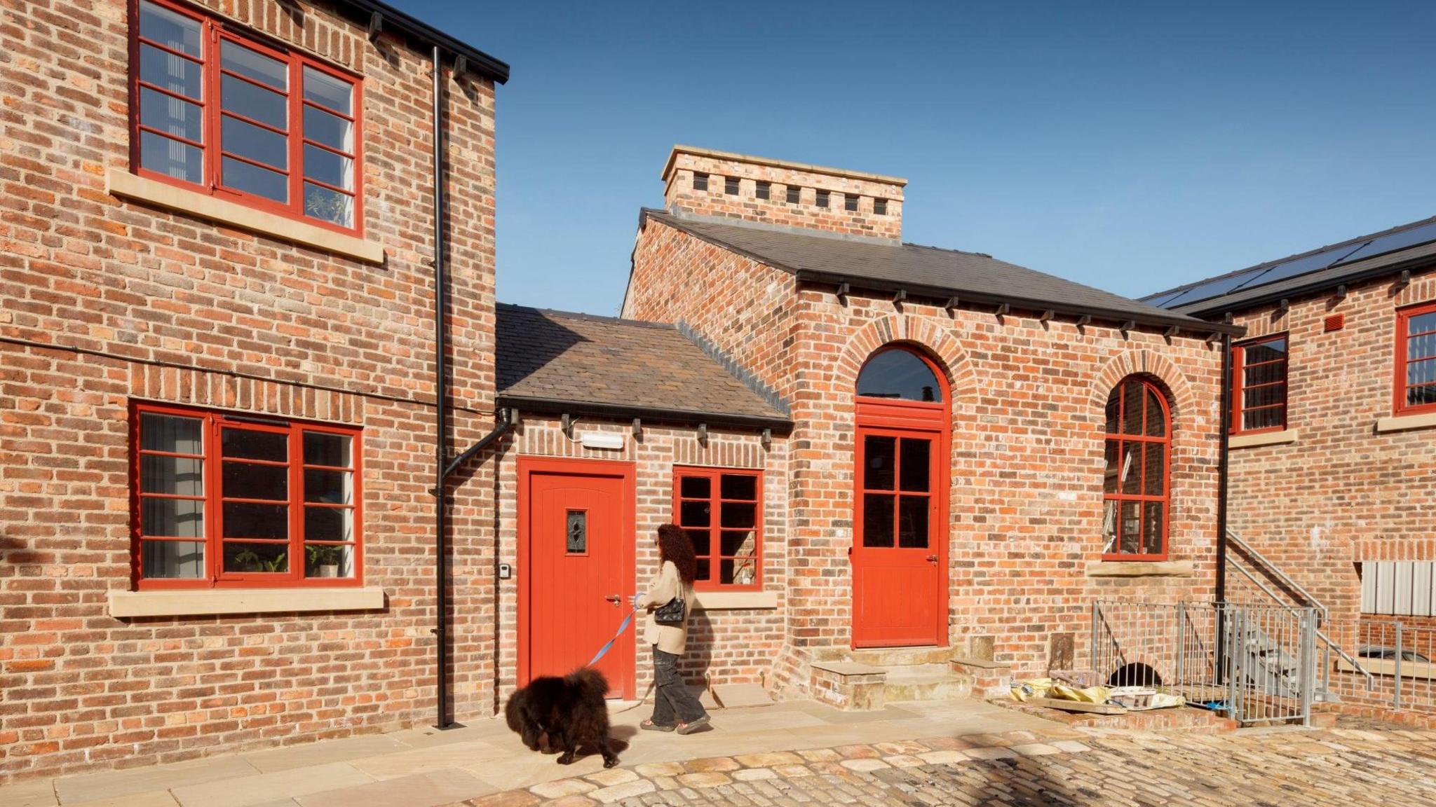 A person walks a large, dark-coloured dog on a lead across a cobbled courtyard. Behind them is a row of well-maintained brick buildings with red-framed windows and doors, showcasing a mix of arched and rectangular architectural features.