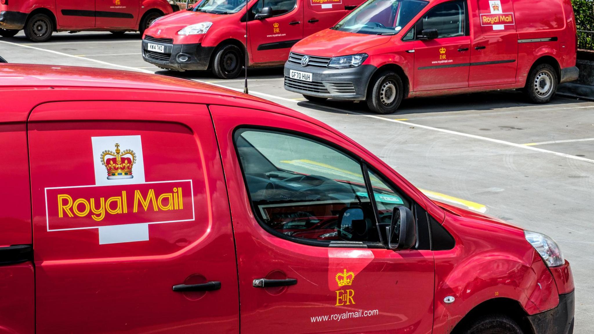 A close-up picture of a red van with the Royal Mail logo and title on the side. It is in a car park with three similar vans beyond.