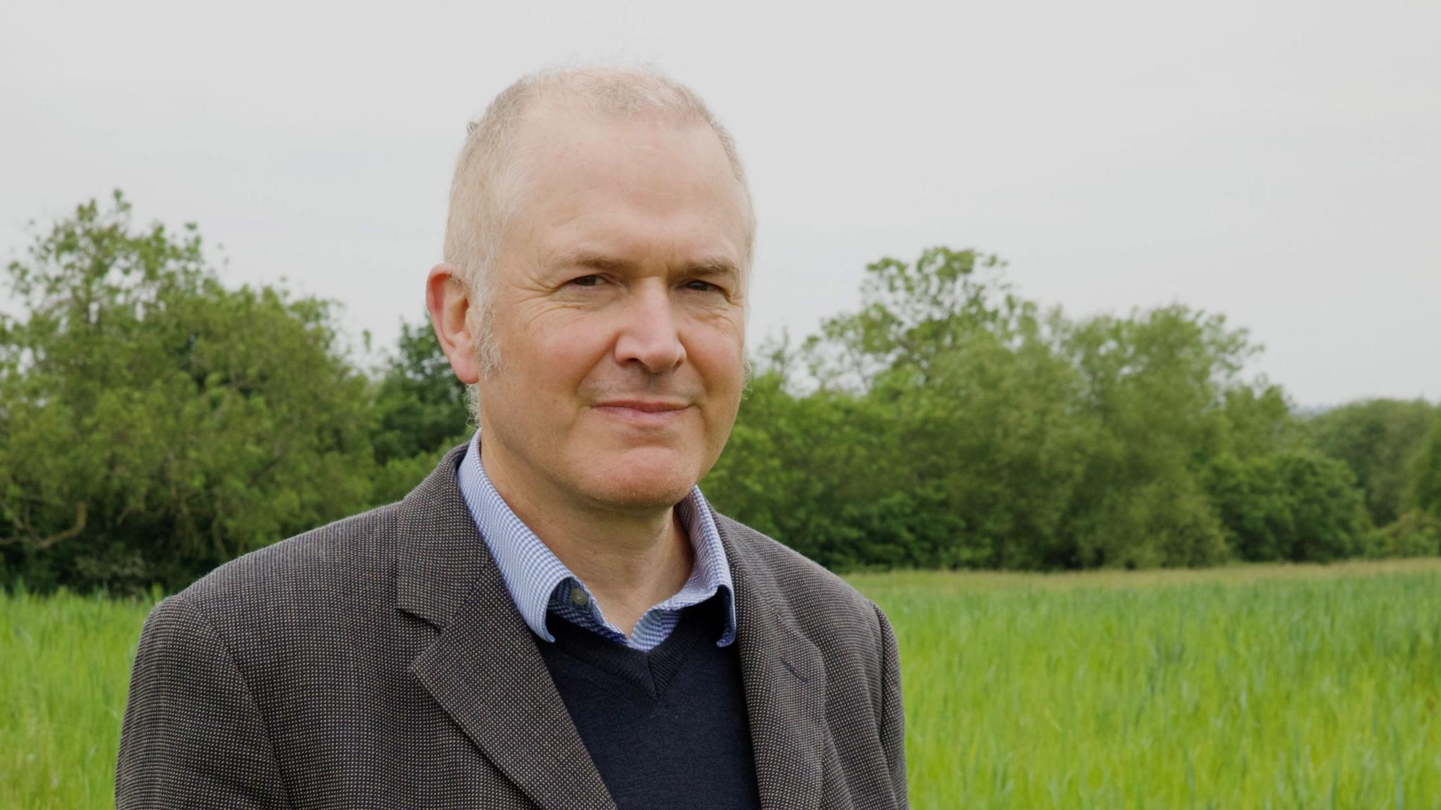 Justin Neal, a white man with closely cropped greying hair who is wearing a brown jacket, a blue shirt and a navy jumper, standing in the empty field where the houses are due to be built, with bushes and hedges behind him.