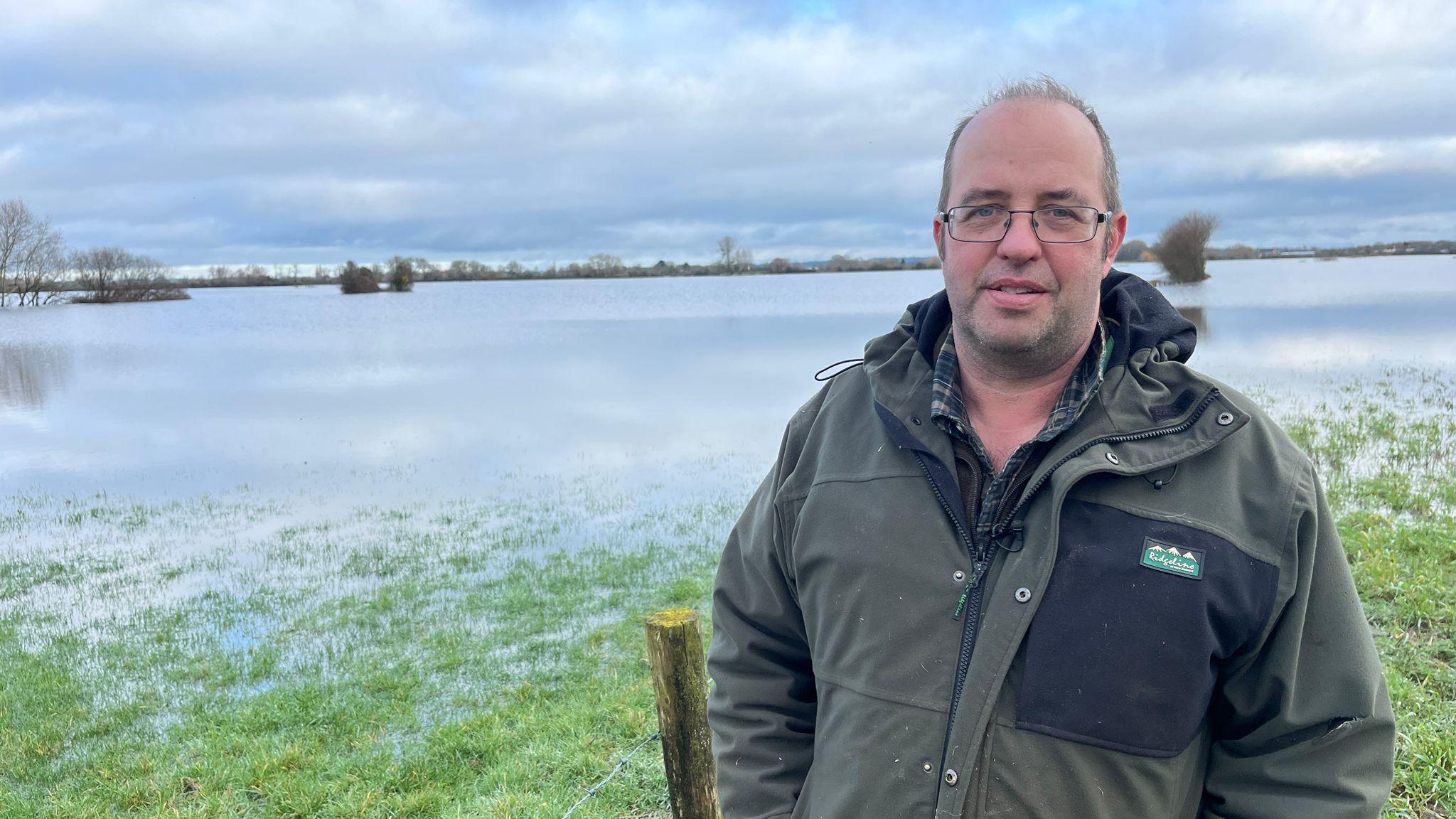 Një burrë i veshur me një xhaketë jeshile dhe këmishë me katrorë qëndron në të djathtë të një fushe me bar e cila është zhytur nën ujin e përmbytjes, skaji i një gardhi mund të shihet pas tij.