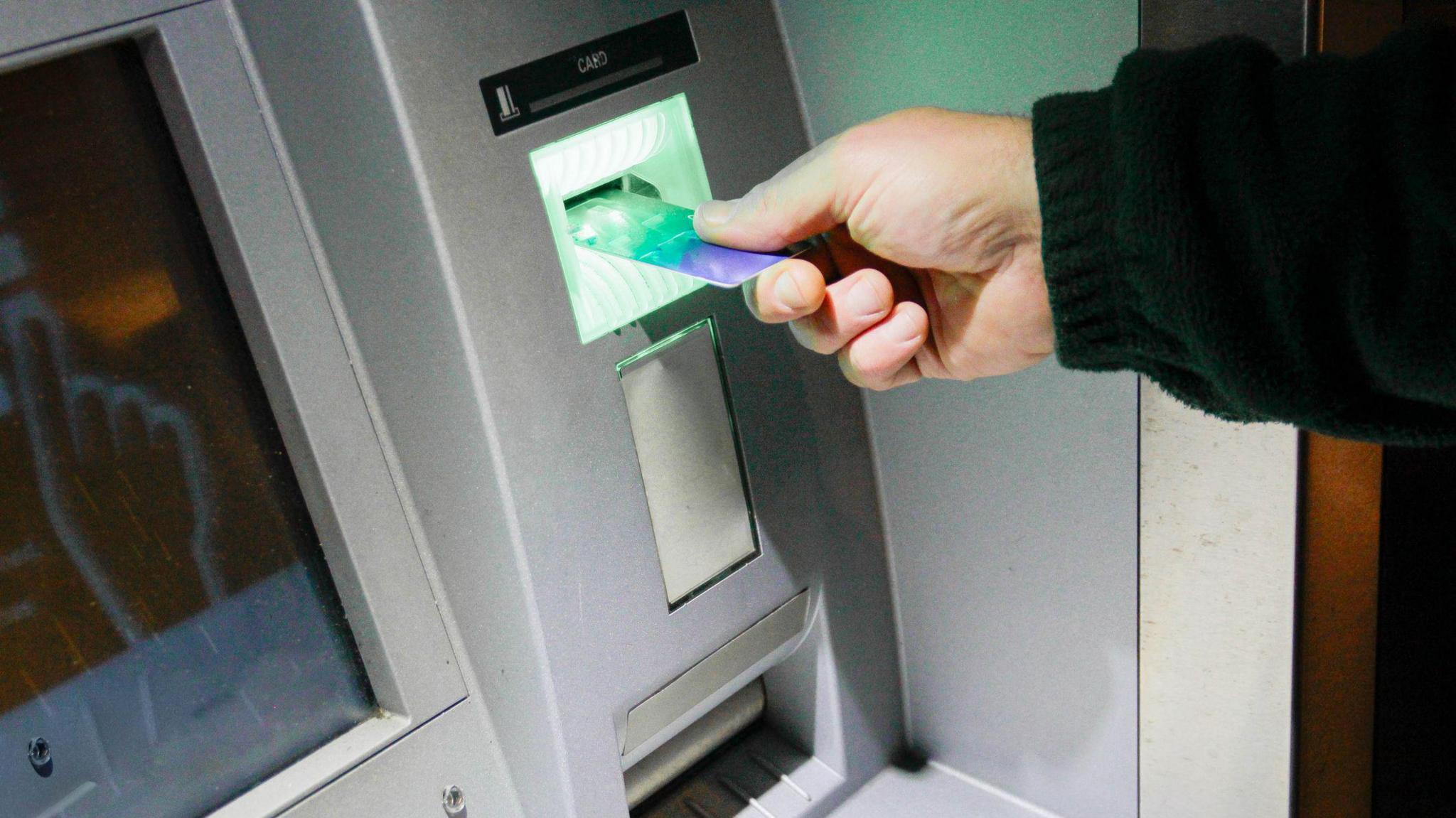 A hand is outstretched holding a credit card, which is being put into a grey cash machine that is mounted into a wall