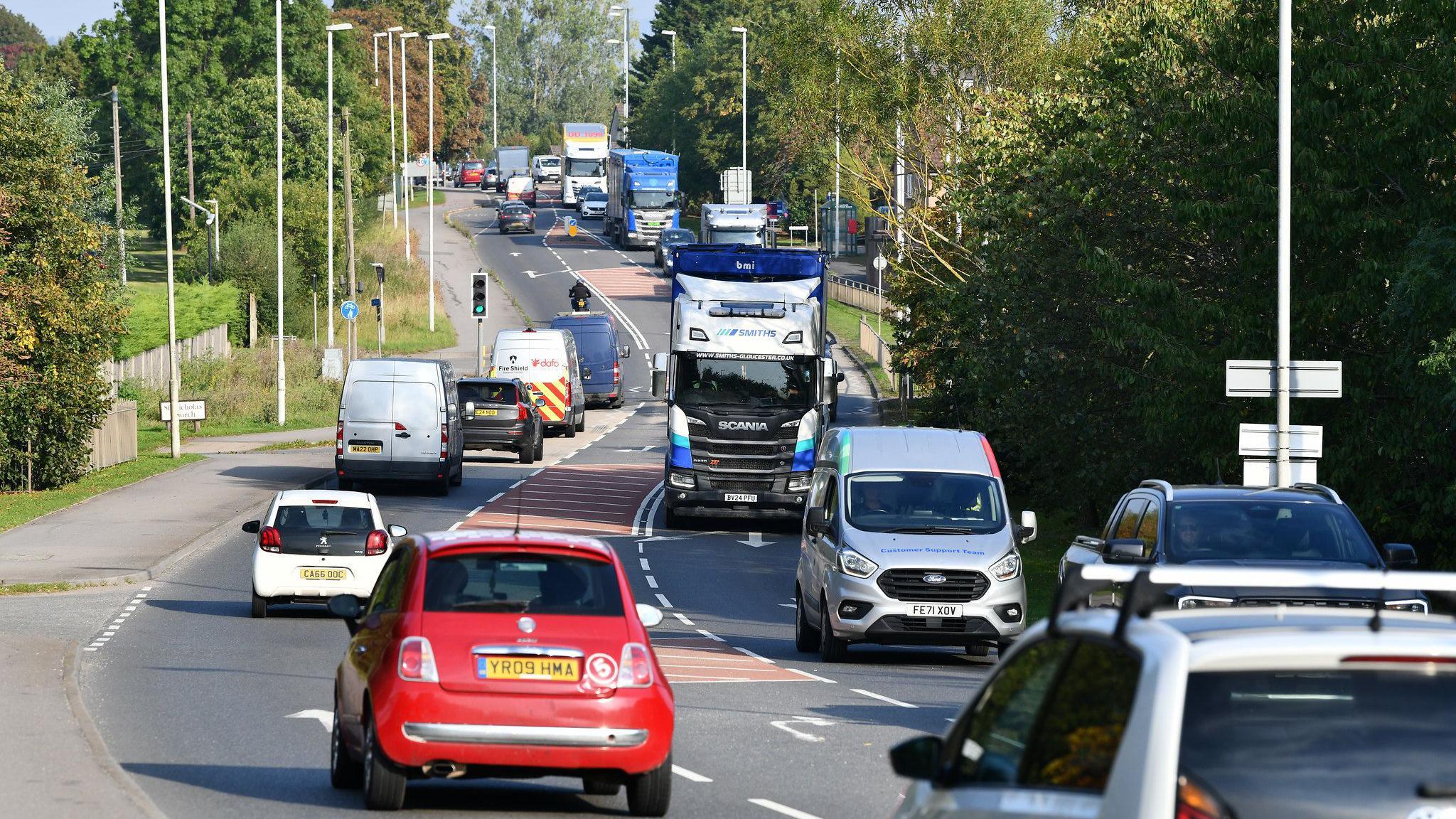 Traffic on the A46 in Ashchurch. It is a slightly curved road with three lanes and trees lining either side. There is a mix of vehicles including cars, vans, and lorries. 