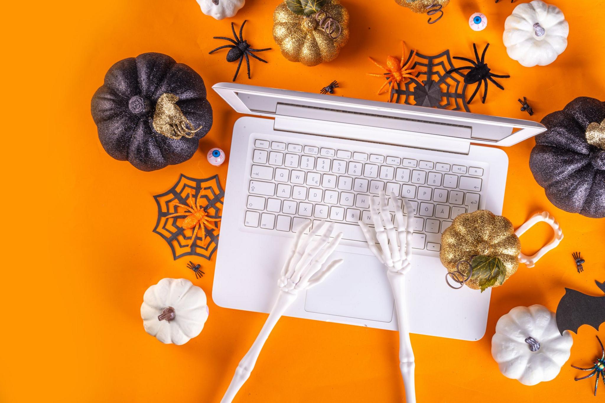 fake skeleton hands typing on a laptop with decorative pumpkins and bats surrounding the laptop