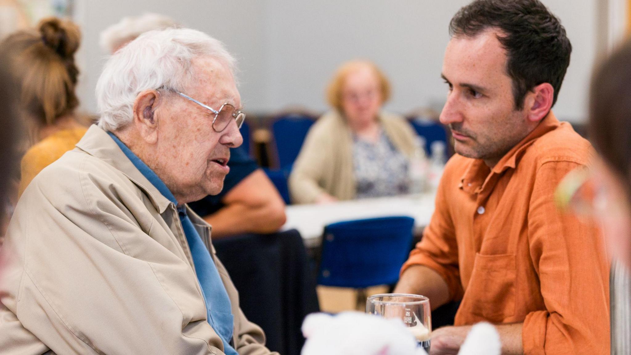 Adam Cooper has short brown hair and is wearing an orange shirt. He is talking to an elderly man with white hair and glasses at Blackhall Community Centre. He is wearing a blue jumper and a beige jacket.