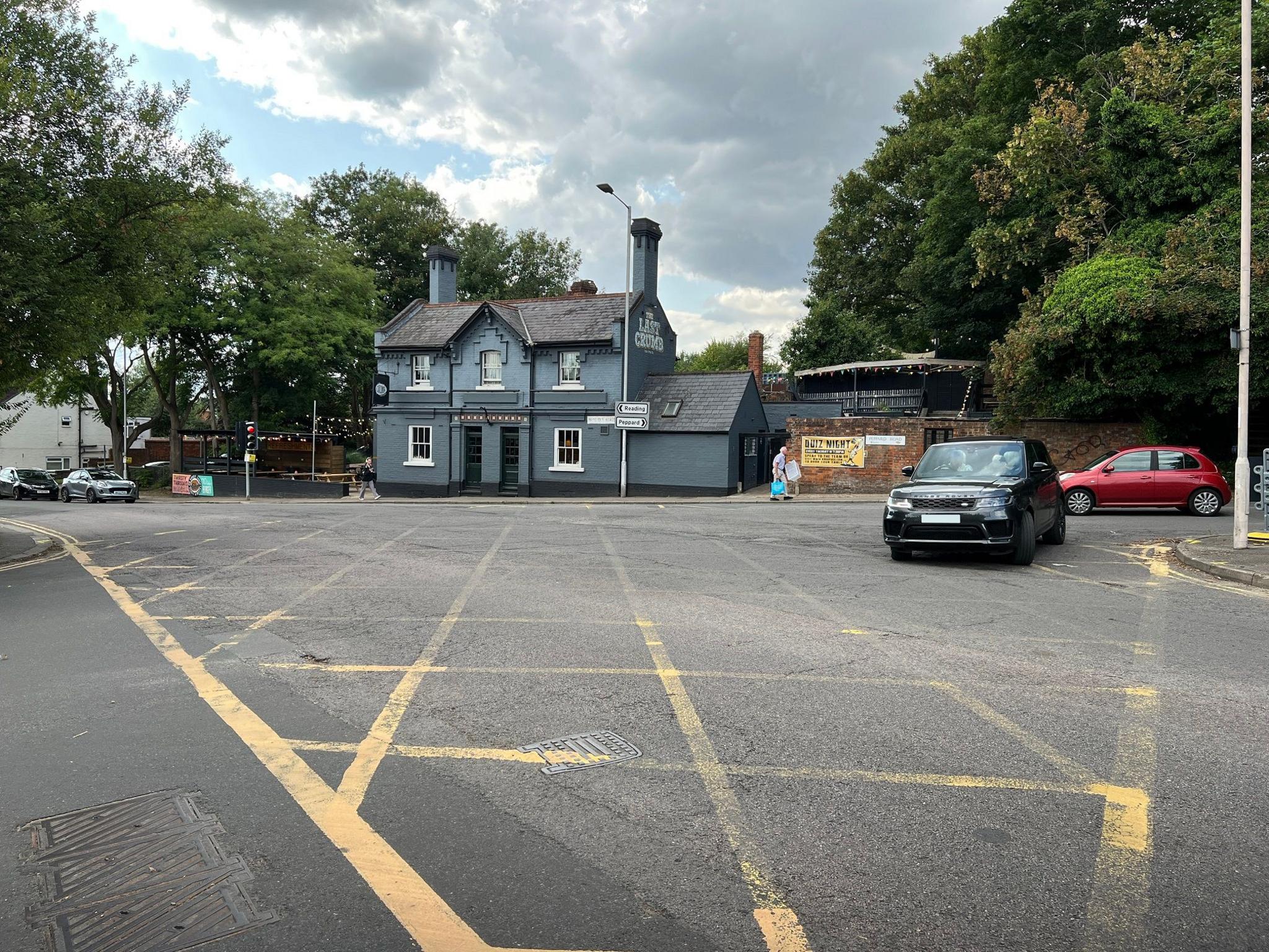 The junction outside the Last Crumb Pub. A yellow-box junction can be seen on the floor and a land rover is making a left turn.