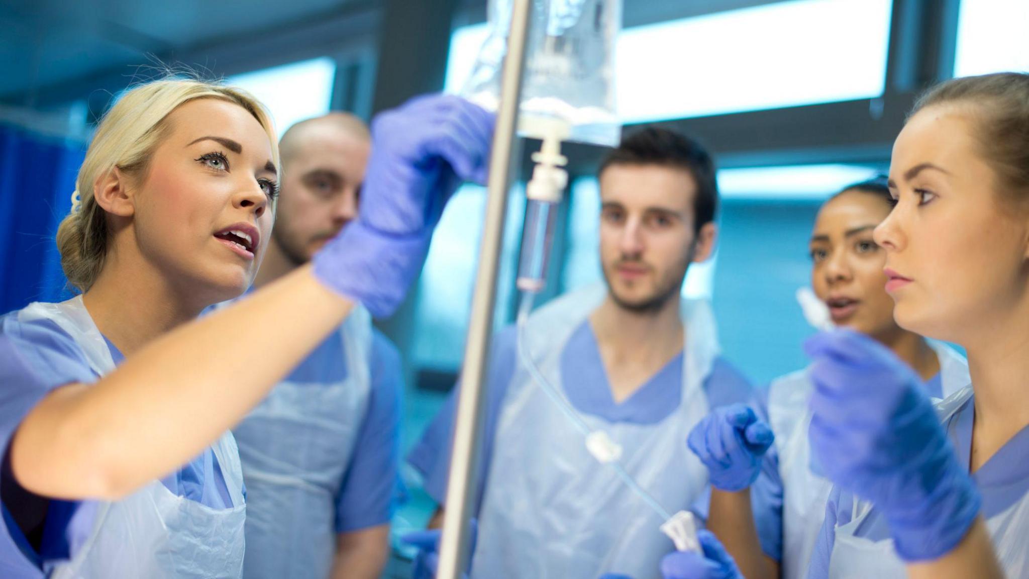 Nurse shows other trainees how to use the IV drip, dressed in blue scrubs and gloves.