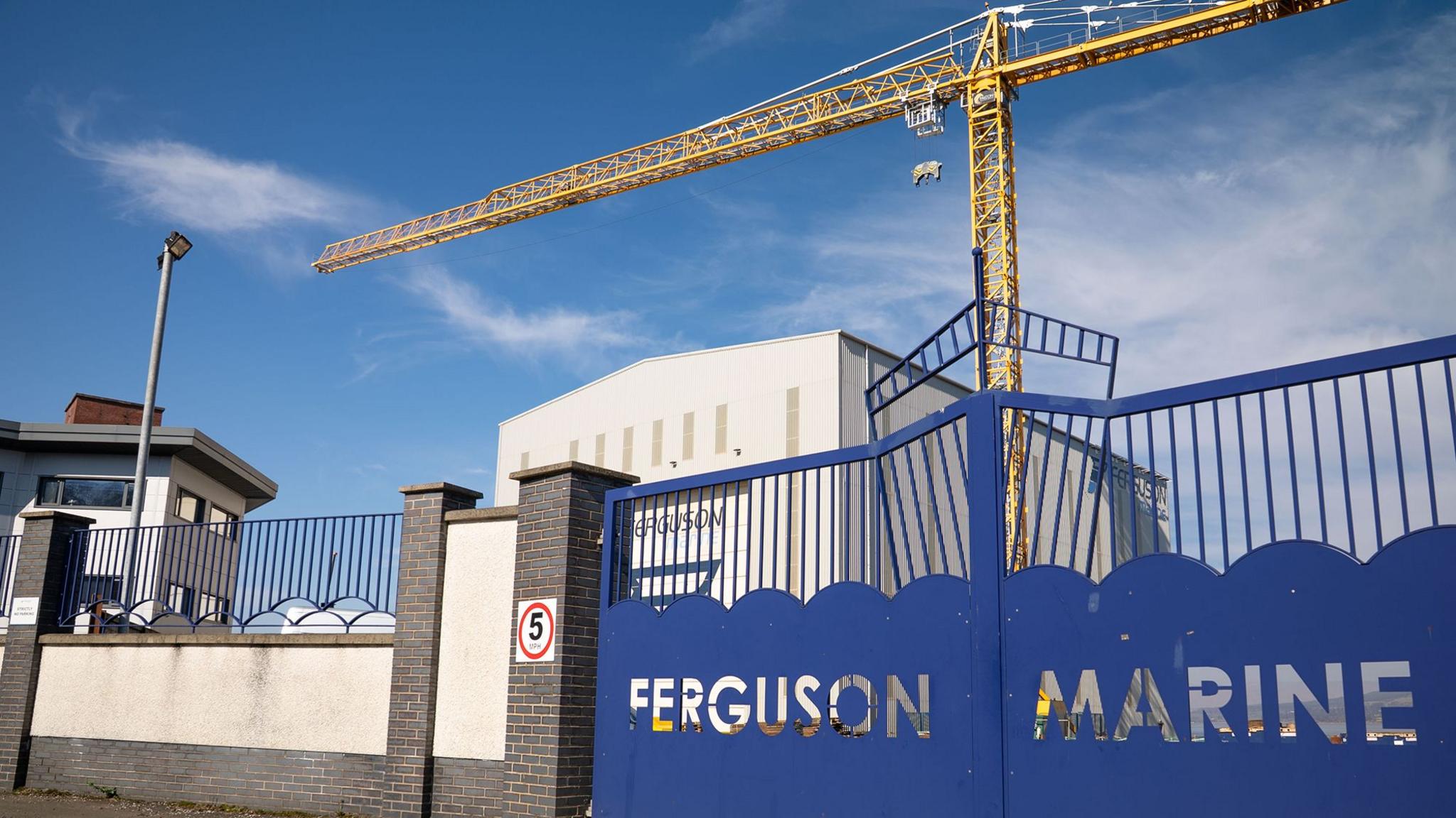 The gates of a shipyard with the words Ferguson Marine cut out of them and a large yellow crane in the background
