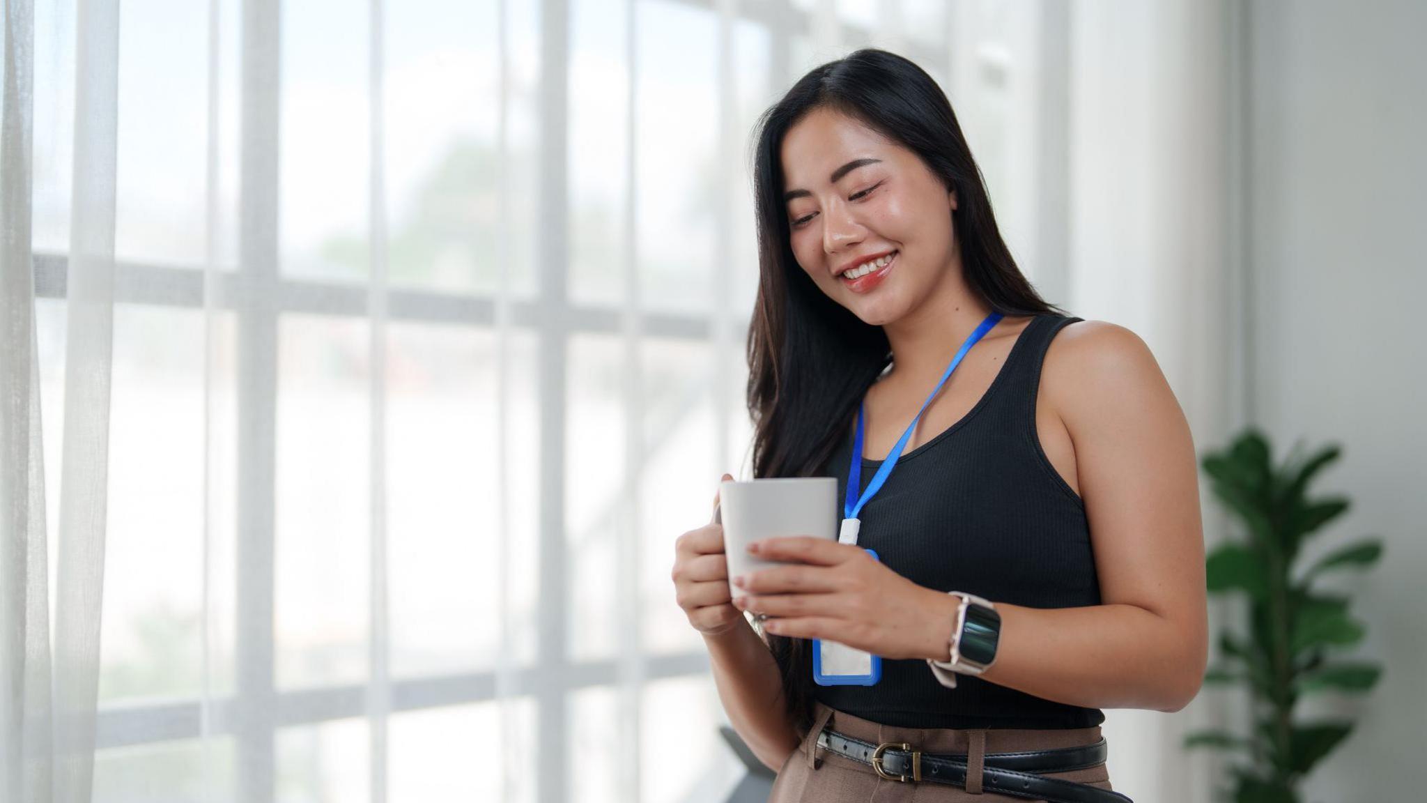 smiling young asian businesswoman standing by the window and drinking coffee during office break