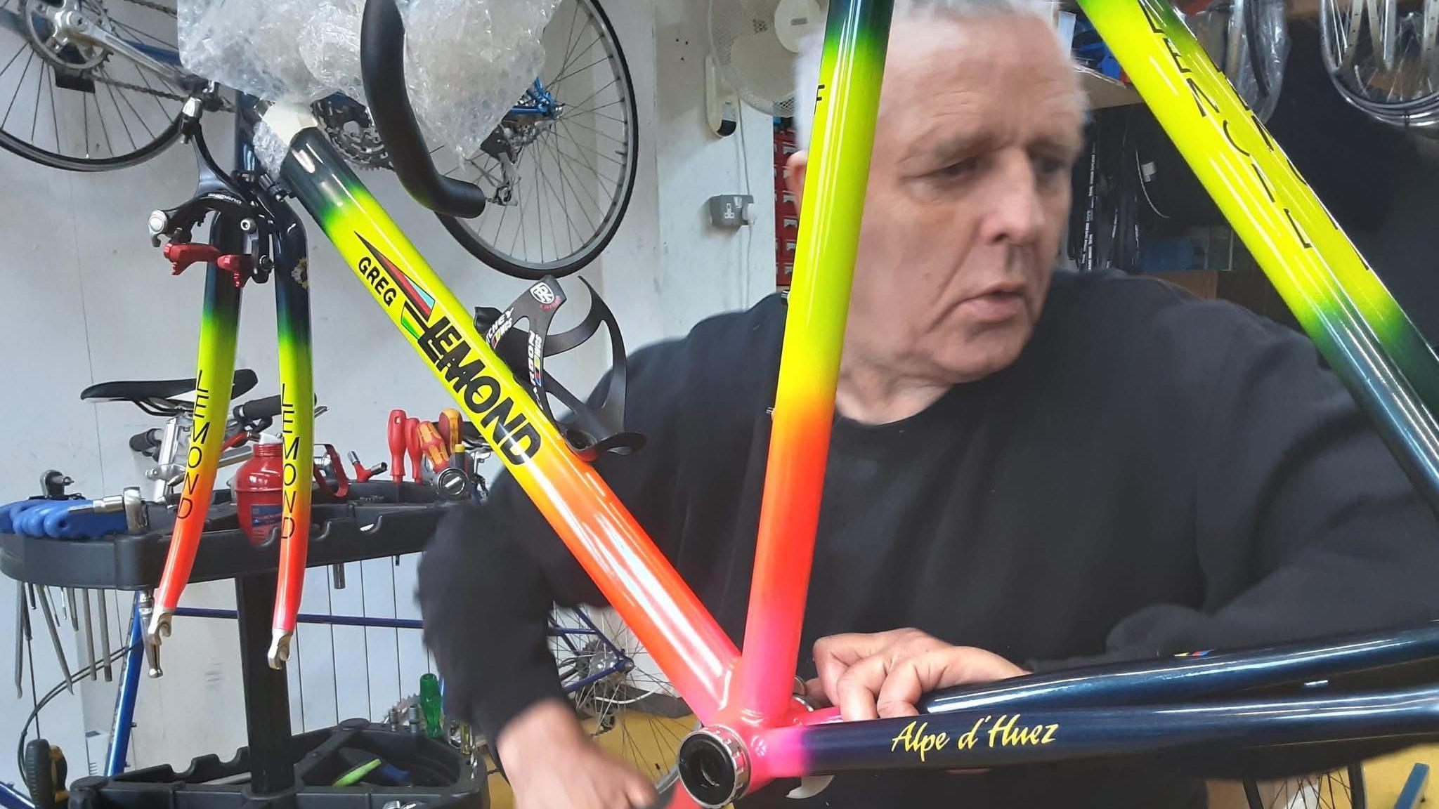 Graham Hicks has grey hair and and a black top is making adjustments to a bike, it just the frame of a striking yellow and red and green cycle, behind him are various tools and machines, with other bikes hanging up.