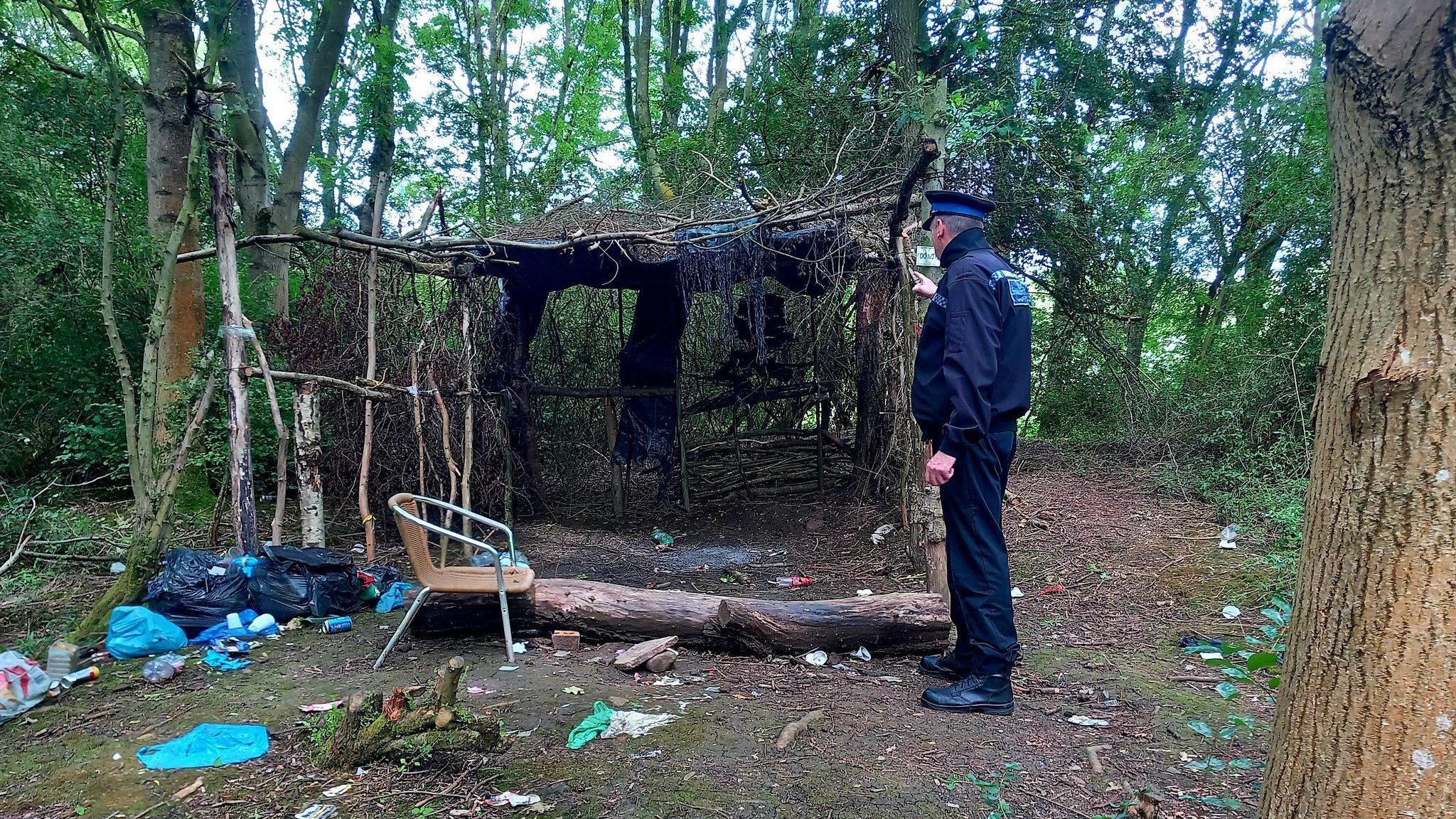 A police office stood in woodland around litter and a wooden shelter made from branches and twigs 