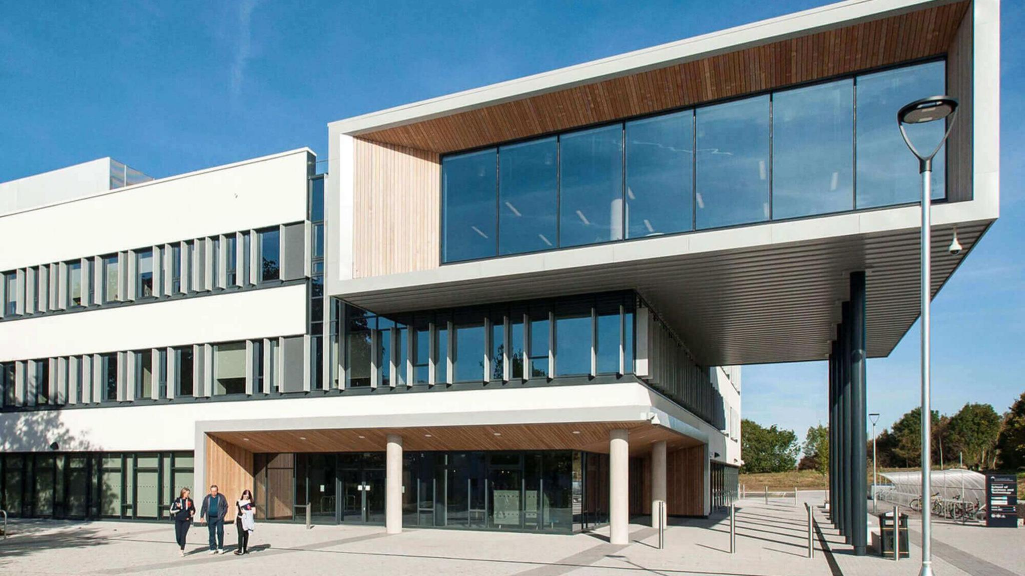 A modern university campus with three storeys and a very large top floor which projects from the rest of the building with large glass windows and wood panelling. It is a sunny day with a clear blue sky and three people are walking away from the building.
