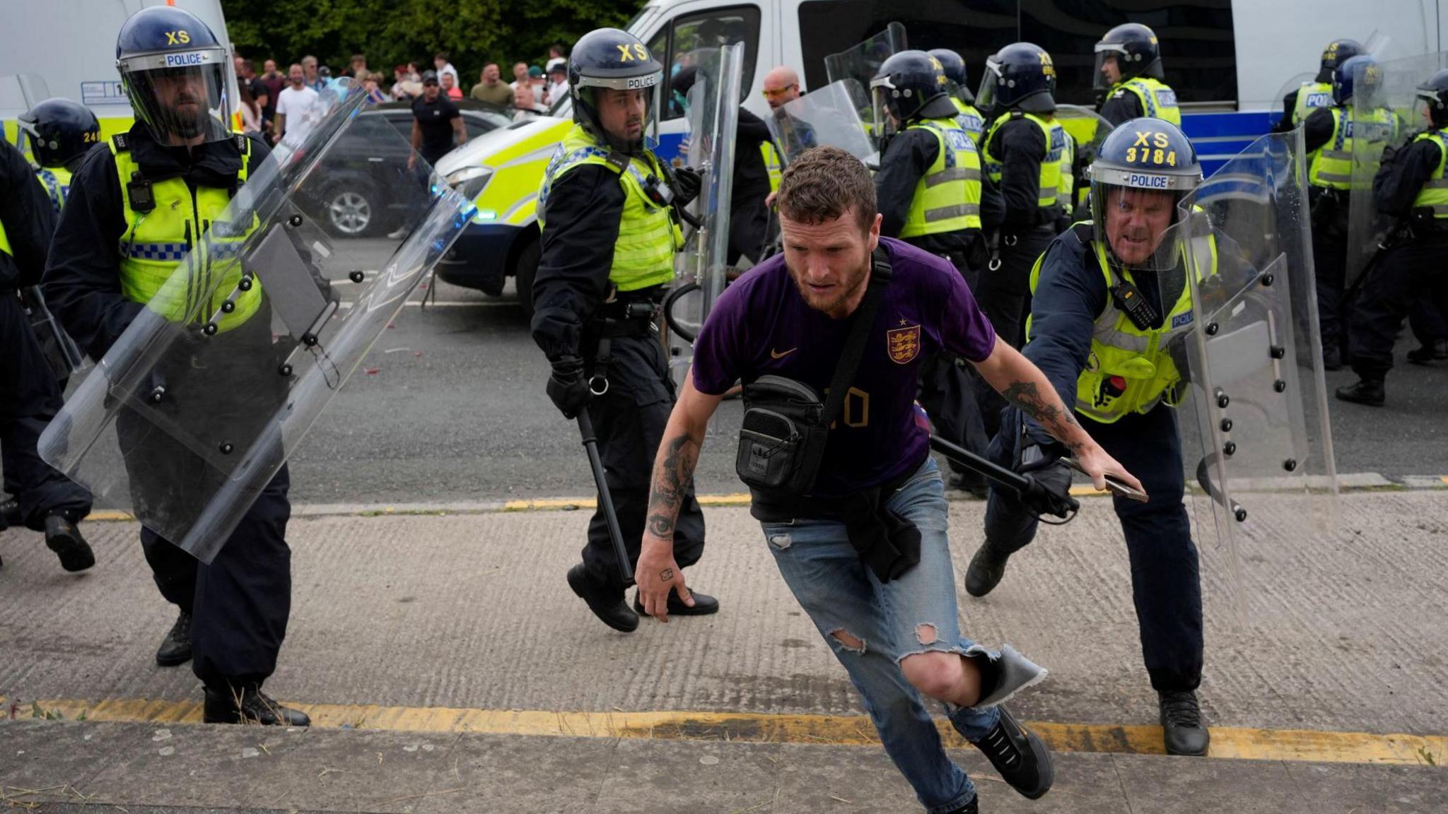 Fishlock wears a purple -tshirt and ripped jeans. He somewhat ducks as he appears to run away from police officers in riot gear. Two police vans can be seen parked in the background.