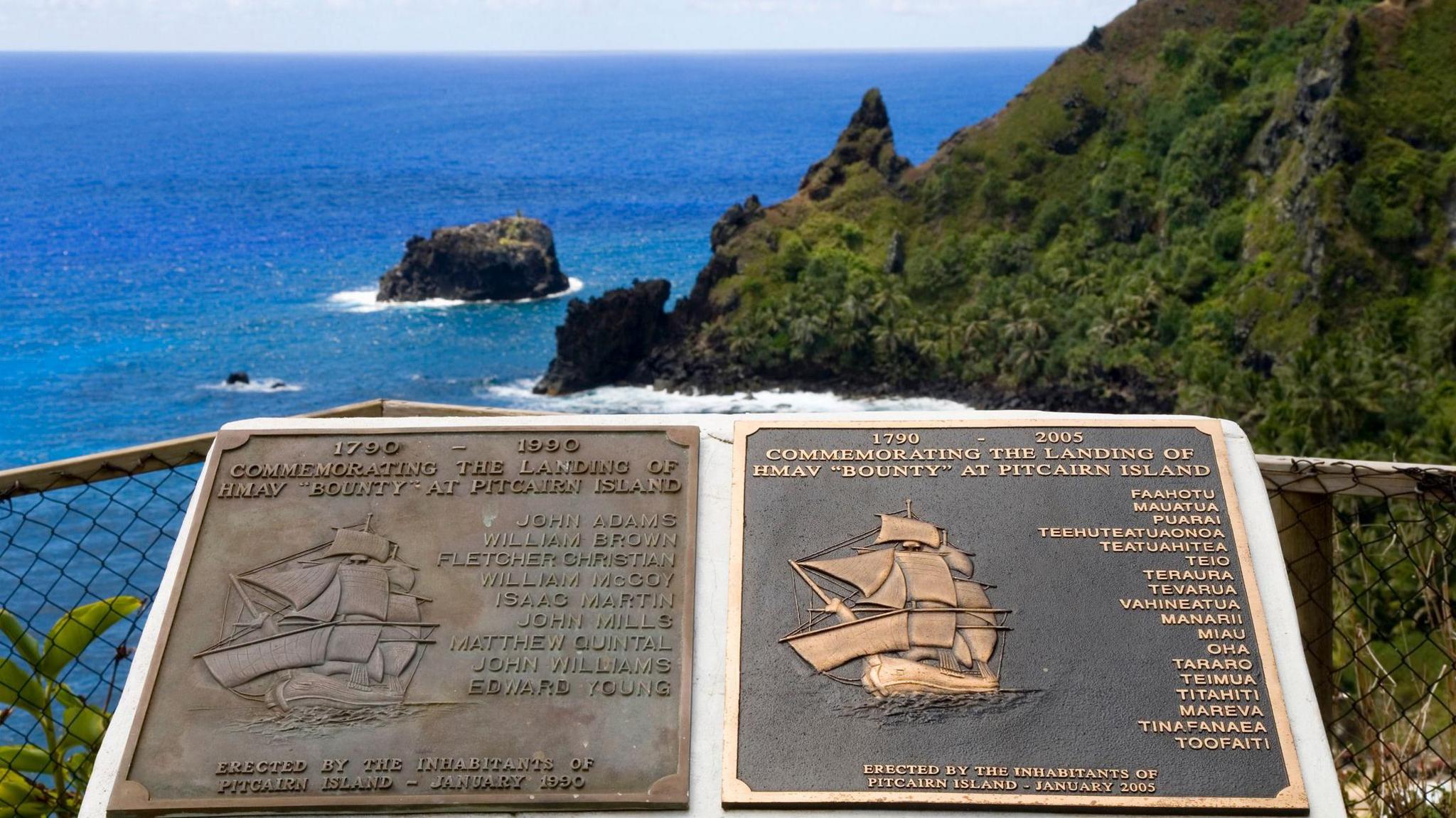 Two plaques commemorating the landing of the Bounty mutineers. They are set in stone on a cliff edge with the sea and a moss-covered cliff in the background.