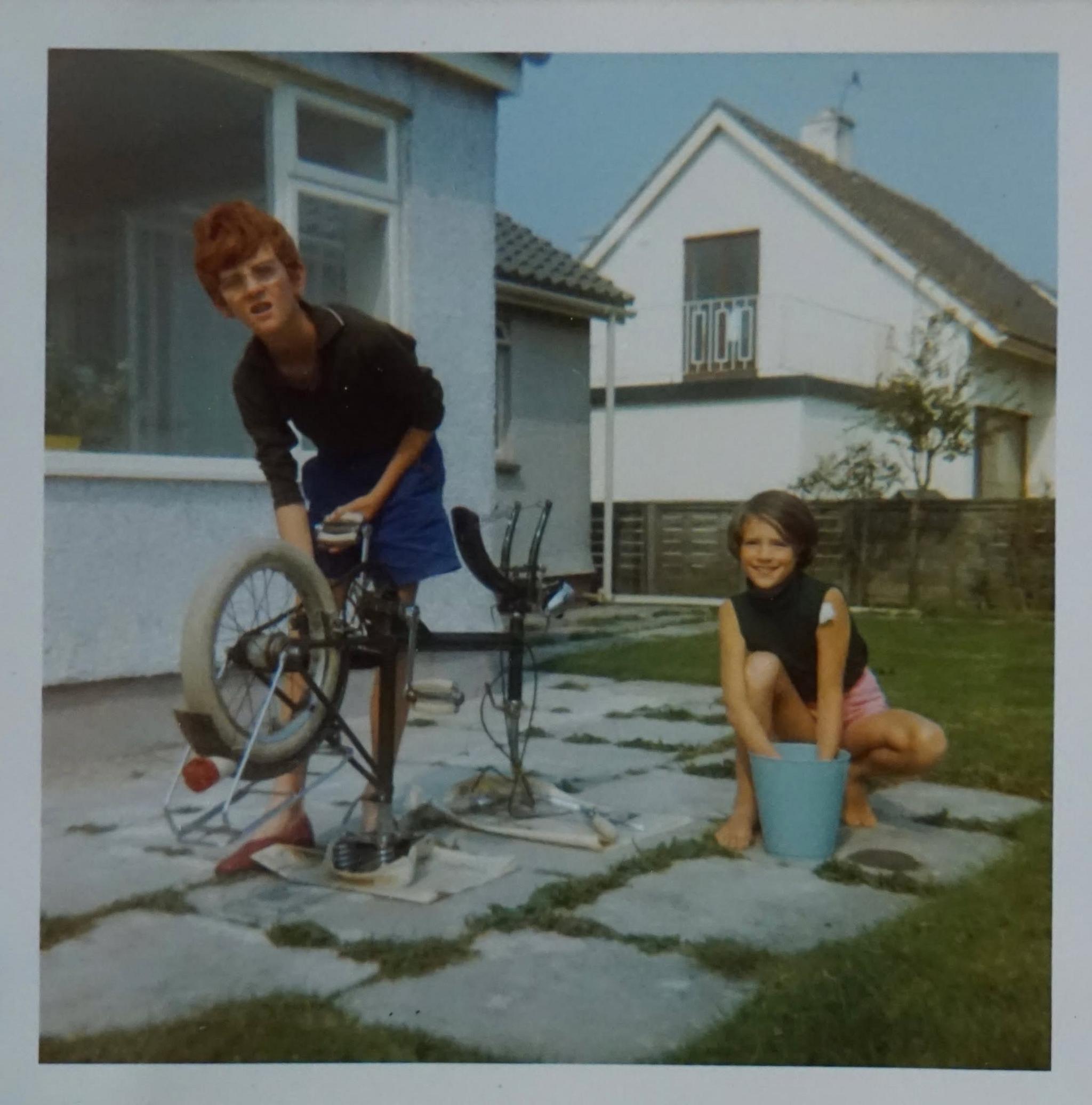 An old photo of two children in a garden - a boy with ginger hair is on the right holding a bicycle - and a girl on the left with short brown hair has her hands in a plant pot.