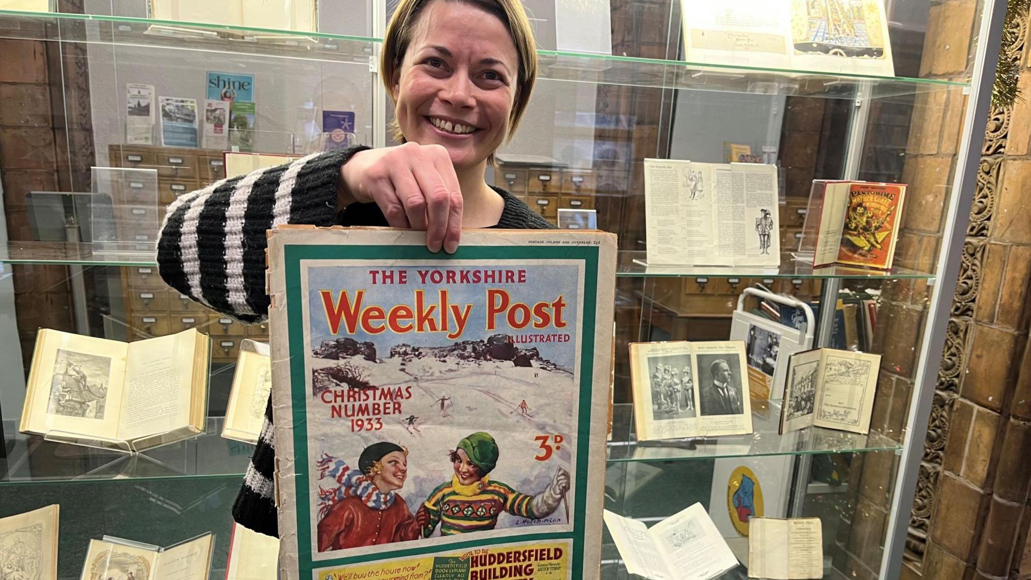 A picture showing senior librarian Rhian Isaac holding a poster of an old front page of a newspaper. The poster is titled The Yorkshire Weekly Post, Christmas Number 1933. On the background is a display cabinet featuring old books.