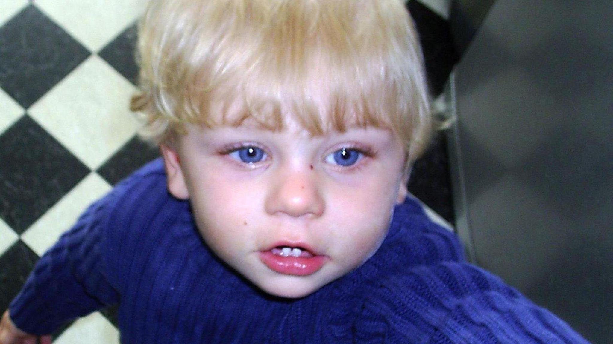 Candid photo of Peter Connelly, a 17-month-old boy. He is blond and blue-eyed and wearing a blue jumper. He is looking to the right of the camera and seems to be reaching for something