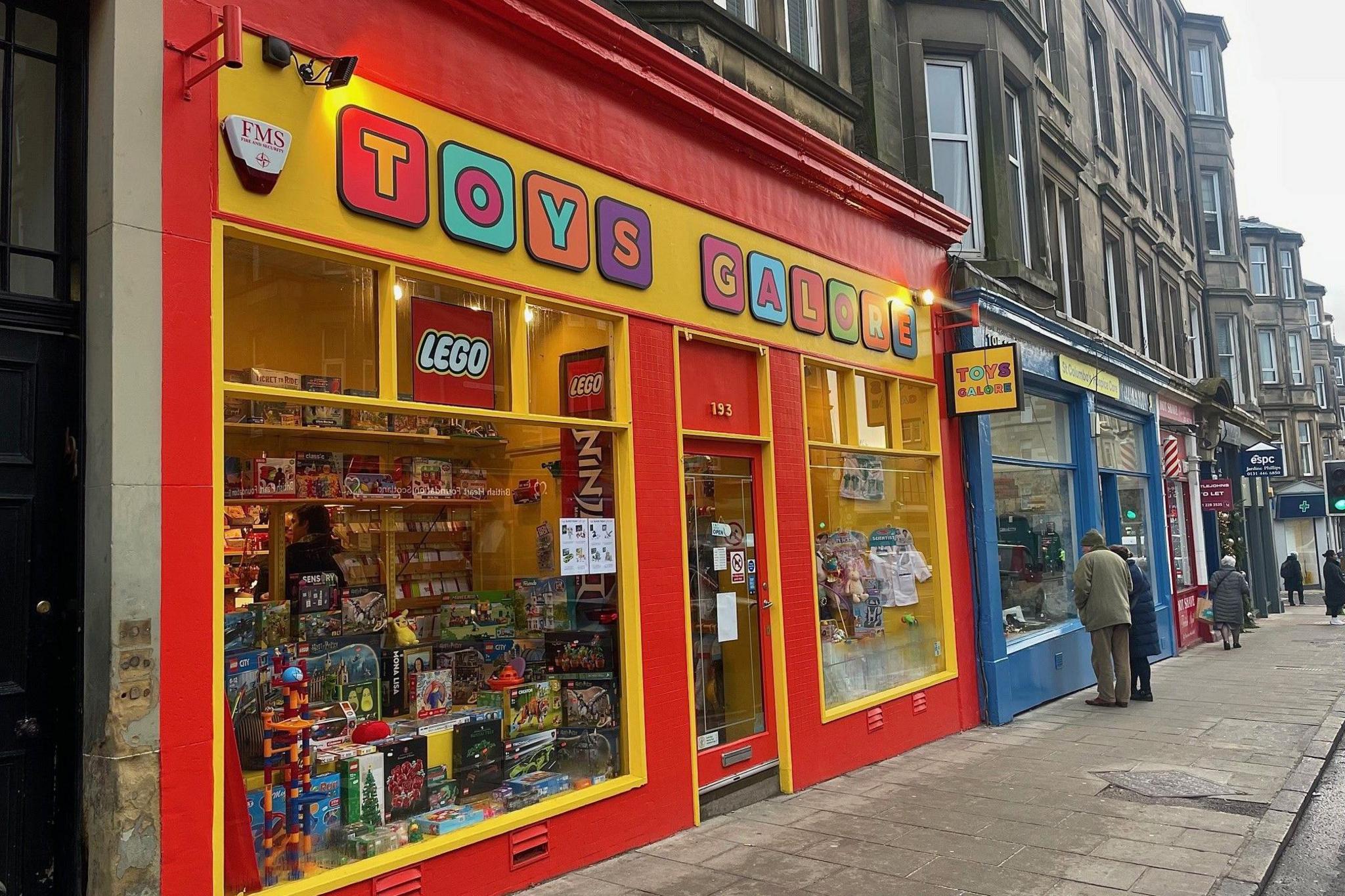 This is the shop front of Toys Galore. It is brightly painted red and yellow and the letters of the shop name are multi-coloured. In the window is a jumble of toys.