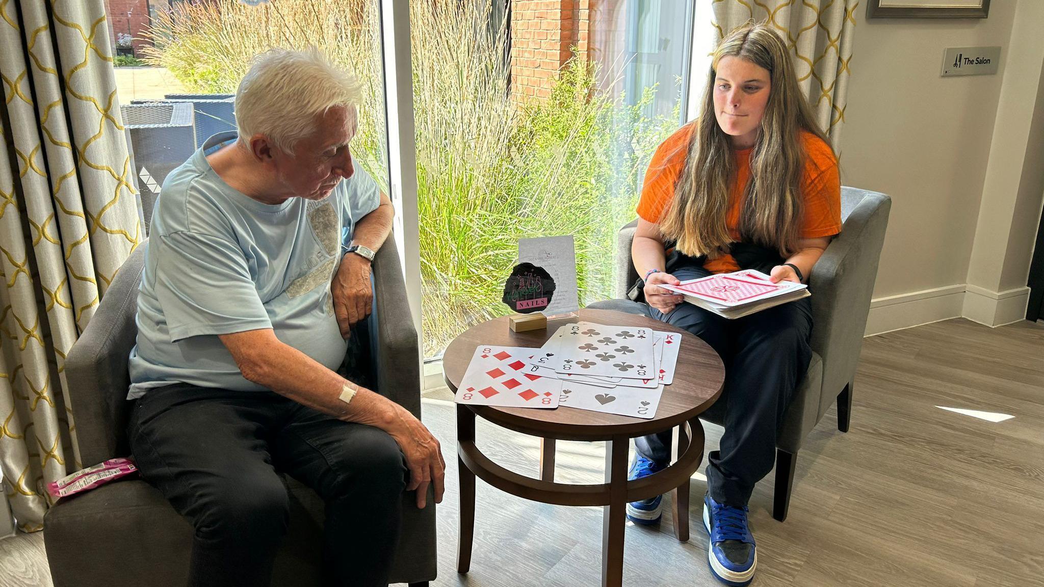 A teenage girl with long brown hair and wearing an orange t-shirt and dark trousers sits at a table with a grey-haired man wearing a light blue t-shirt and dark trousers. The girl is laying out large playing cards on a small wooden table.