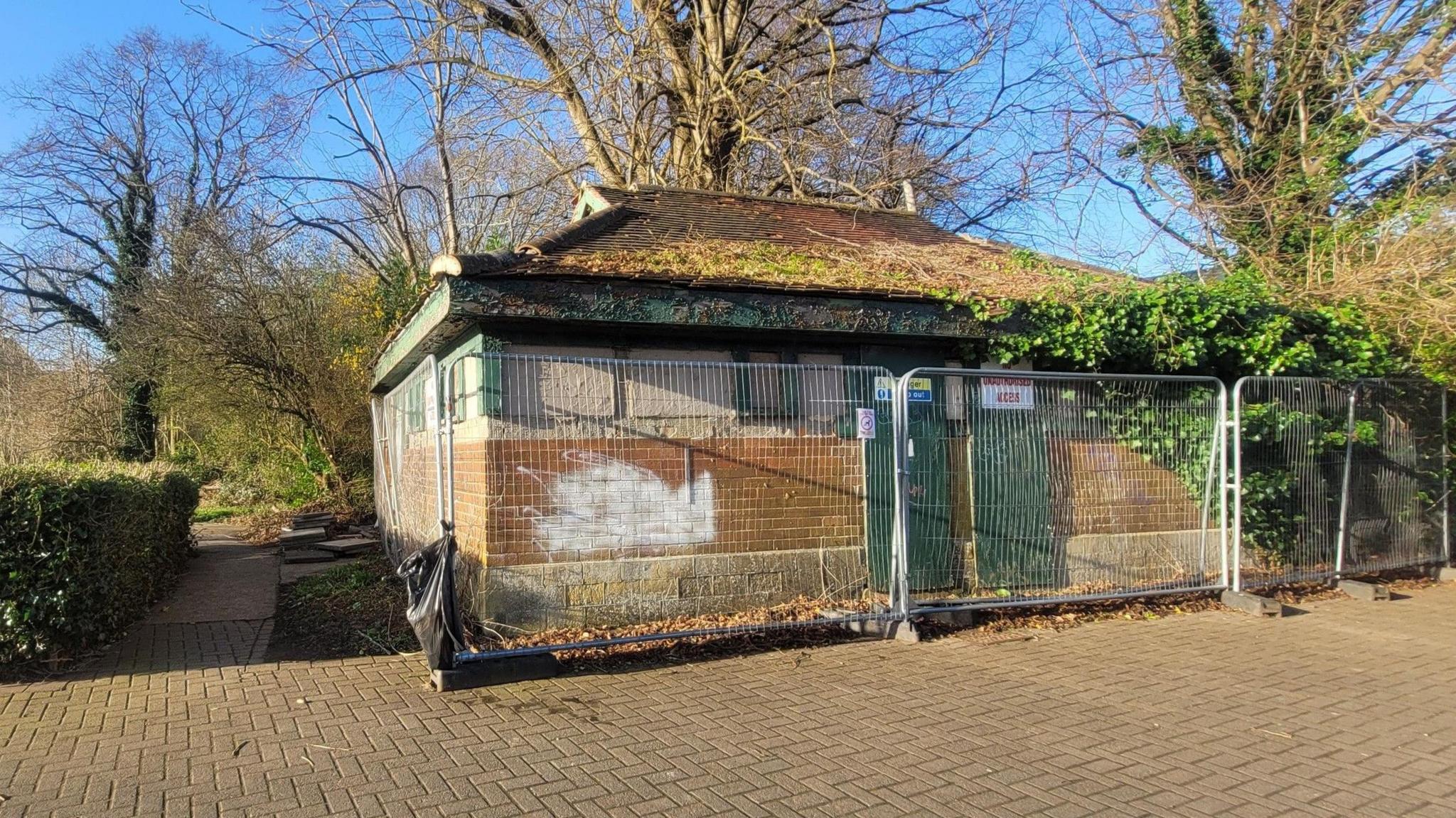 The fenced off toilet block pictured in March 2025. It is a one-storey brick house with green doors and matching window seals. It is surrounded by a metal fence.