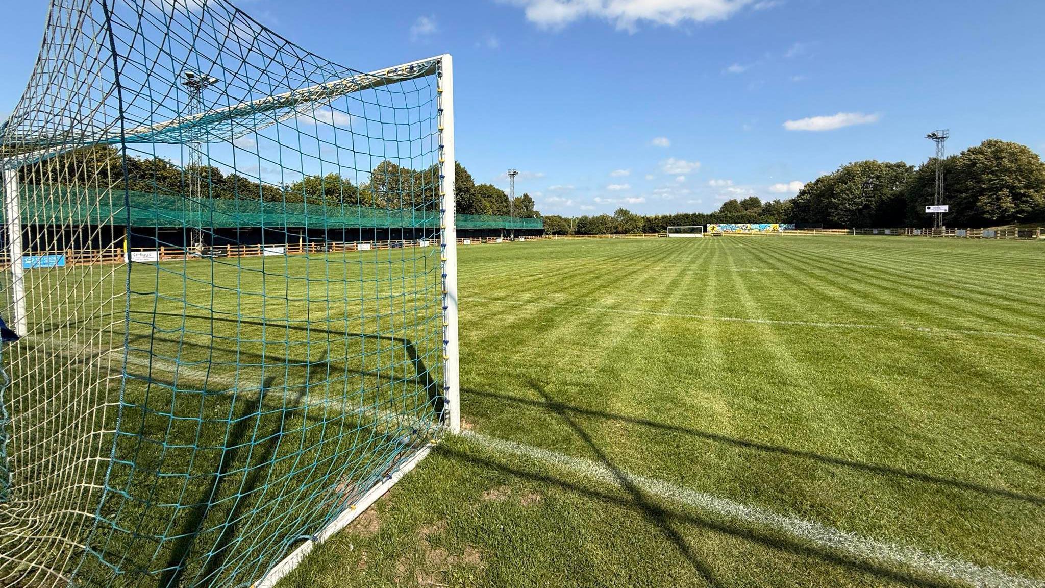 A stock image of a football pitch with green grass and a goal with blue and white netting. There are stands and trees in the far distance.