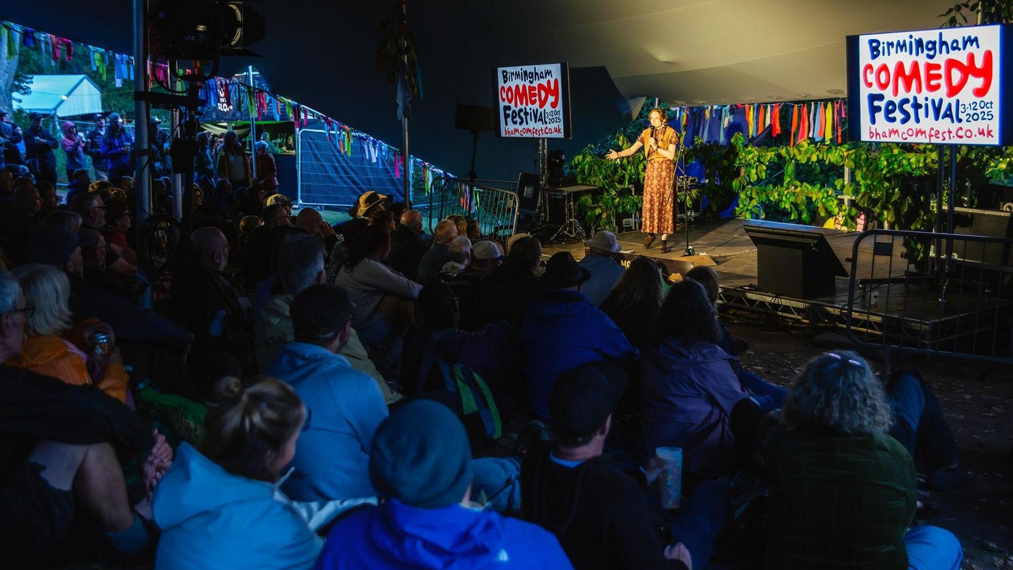 Comedian Rachel Baker wearing a brown flowery dress is flanked by two screens which read Birmingham Comedy Festival.
