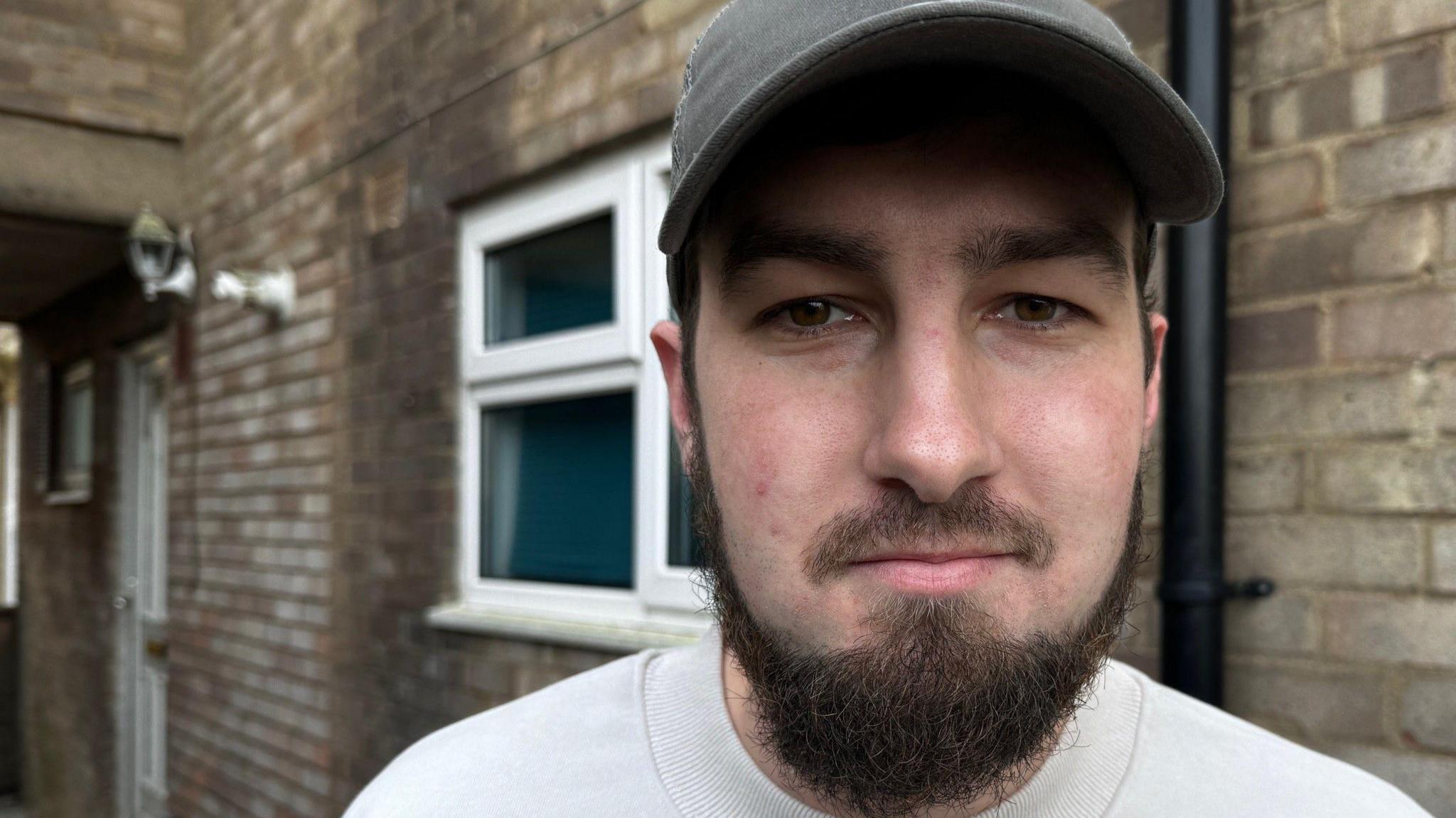 A man with a bear, wearing a baseball cap, wearing a cream jumper, staring at the camera. There is a house behind him