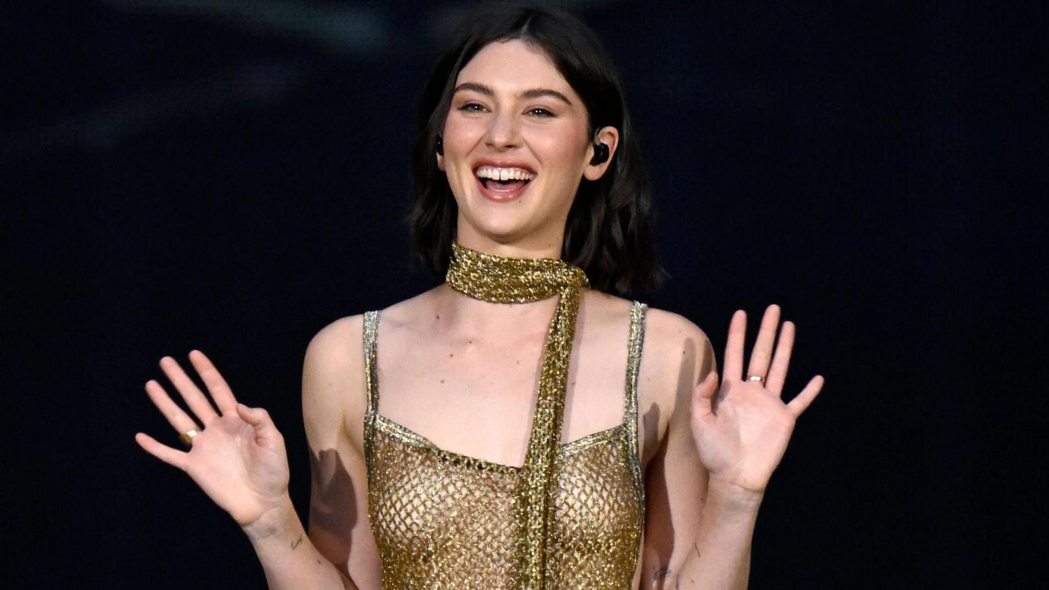 Gracie Abrams on stage during Taylor Swift's Eras tour. Gracie has shoulder length brown hair and brown eyes. She smiles, both hands raised with her palms facing the camera. She wears a crochet gold dress with a matching scarf. 