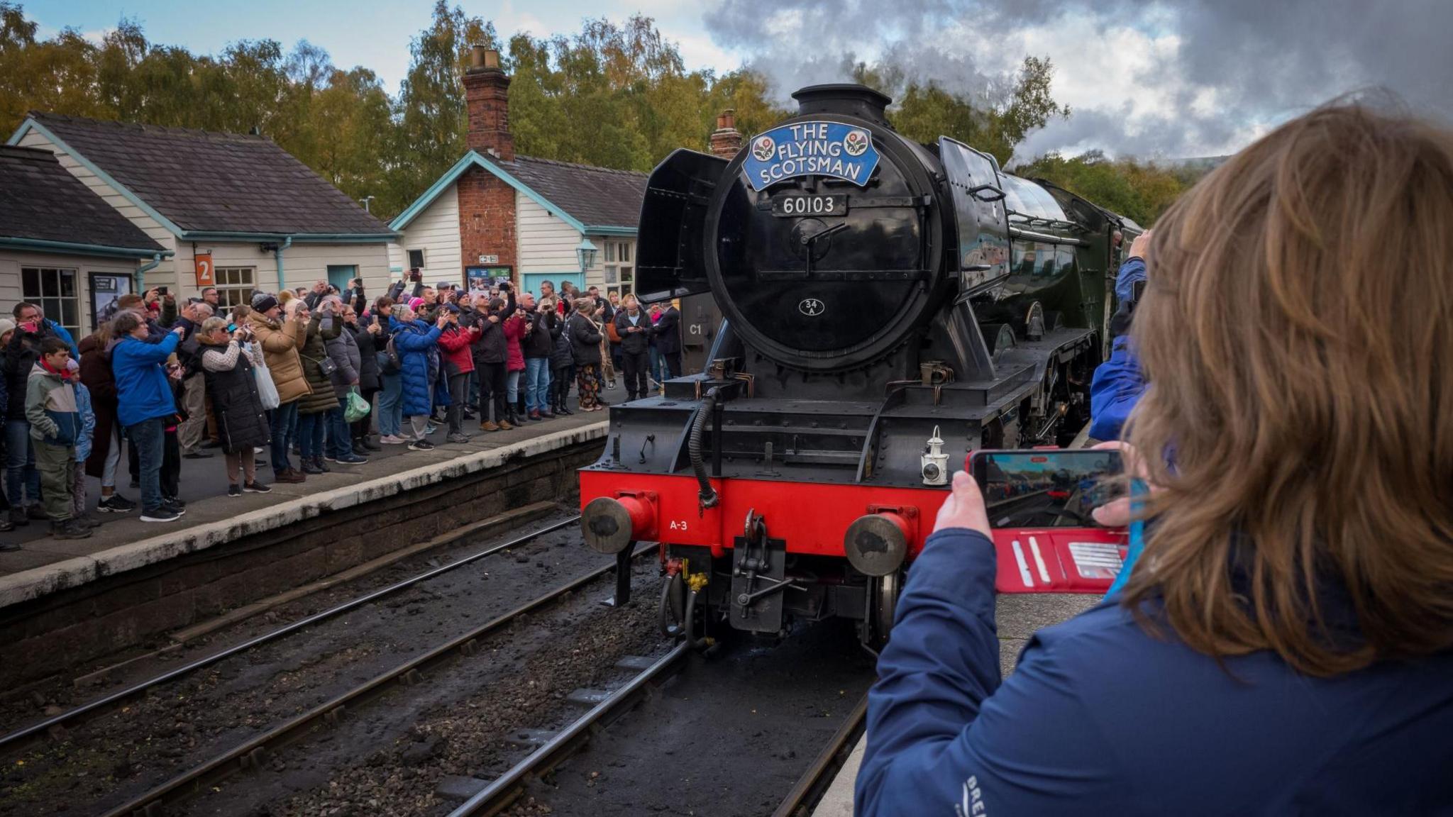 A crowd of people, including families and railway enthusiasts, gather on a station platform to admire and photograph the historic steam locomotive "The Flying Scotsman," numbered 60103.
