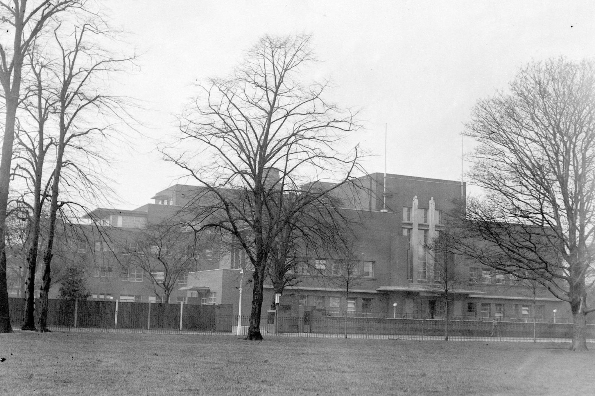 A black and white photo of a large building, seen from a distance across a park. In front of the building, there is an area of grass and a row of leafless trees, followed by a railing and a fence. The building is multiple storeys tall and has a grand, Art Deco style, with windows and a brick design.