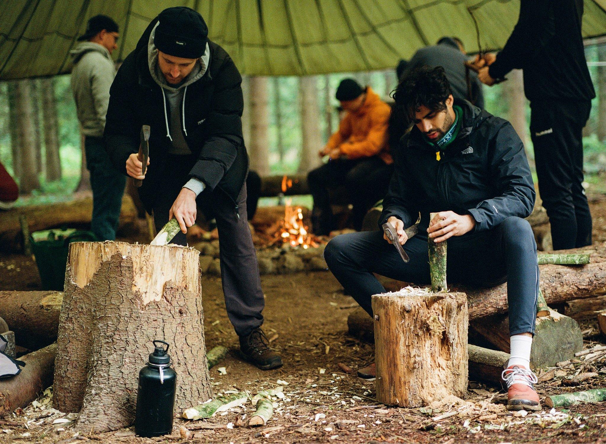 Two men shape wood ontop of tree stumps under a canopy