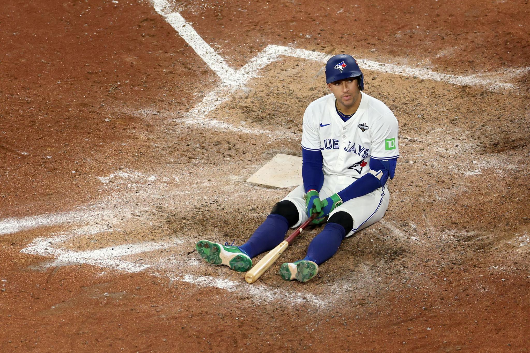 A baseball player in a white Blue Jays uniform sits on the dirt near the home plate, legs extended forward. The player holds a bat resting across the legs, with green gloves visible. White chalk lines outline the batter’s box.