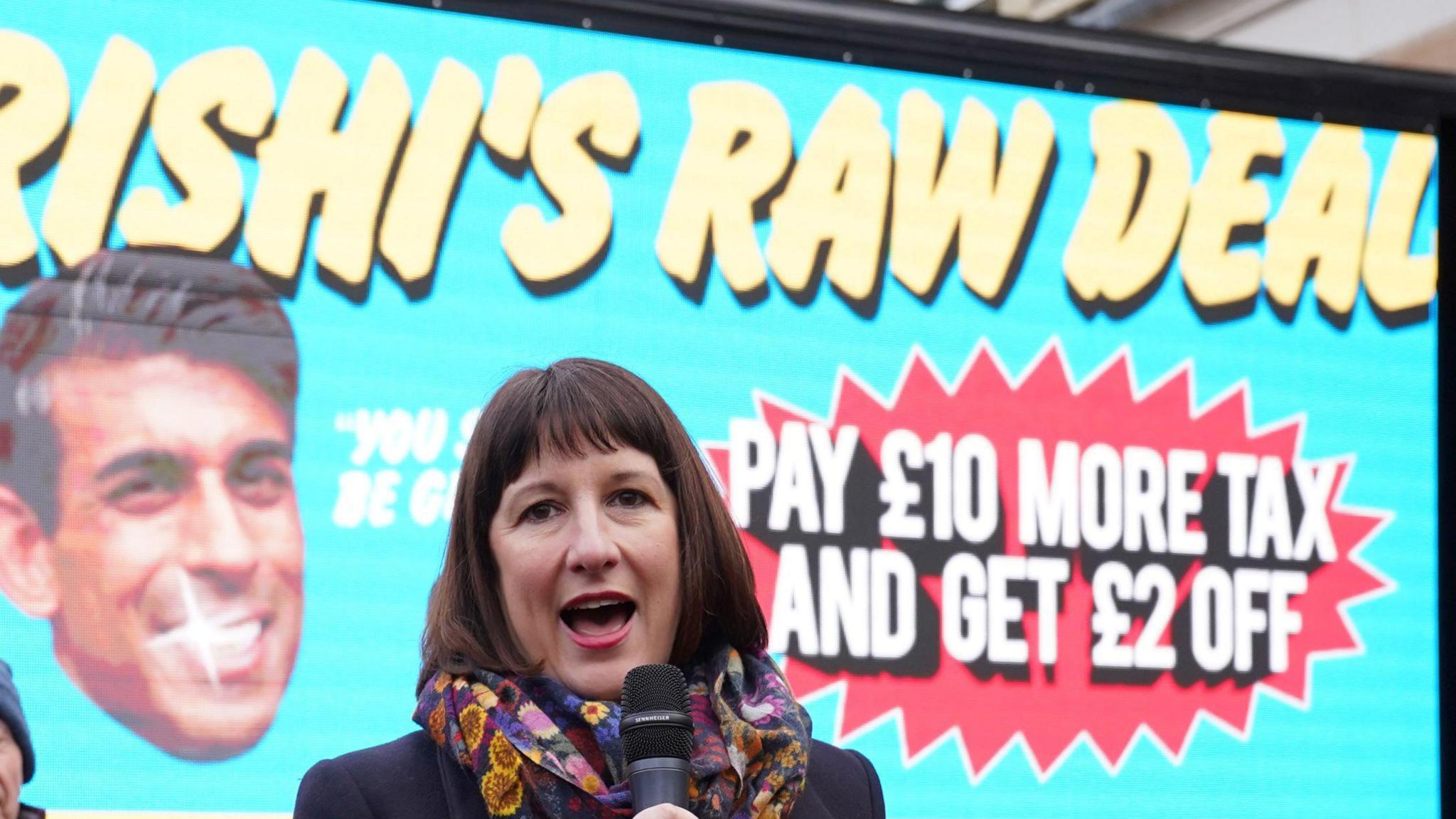 Rachel Reeves, during a visit to Wellingborough, North Northamptonshire, to unveil Labour's poster campaign of what it calls "Rishi's raw deal" for taxpayers ahead of the reduction in national insurance contributions - she has a microphone in hand a multi coloured scarf, with a banner behind her saying RISHI'S RAW DEAL in bigger letters and a photo of Rishi Sunak grinning 