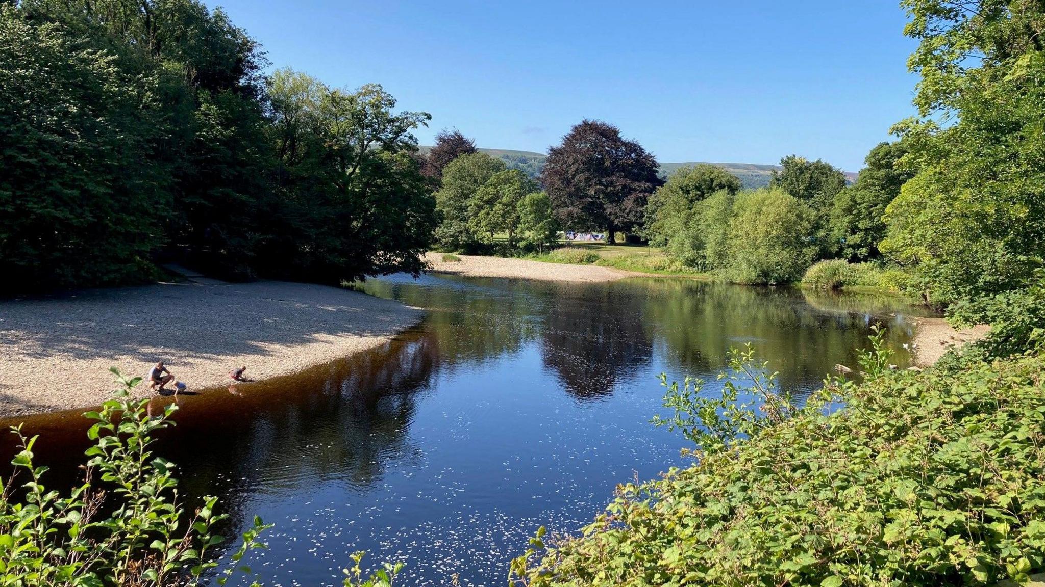 A man and two children play on a visible bank of grey/brown pebbles on a stretch of the River Wharfe.