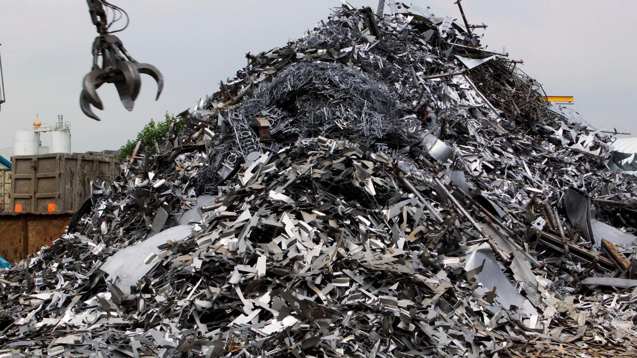 A huge pile of silver scrap metal, with a metal crane claw coming down towards it, to grab it.