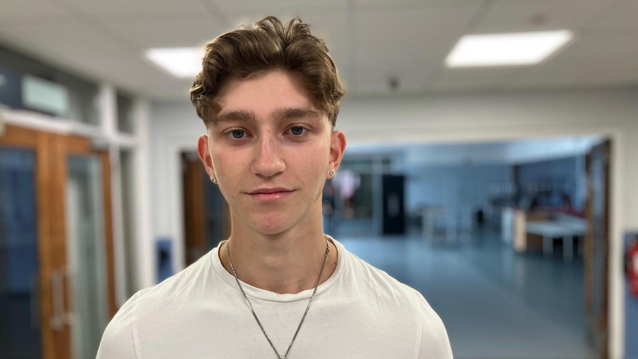 A teenager wearing a white shirt and thin chain necklace with a pendant.