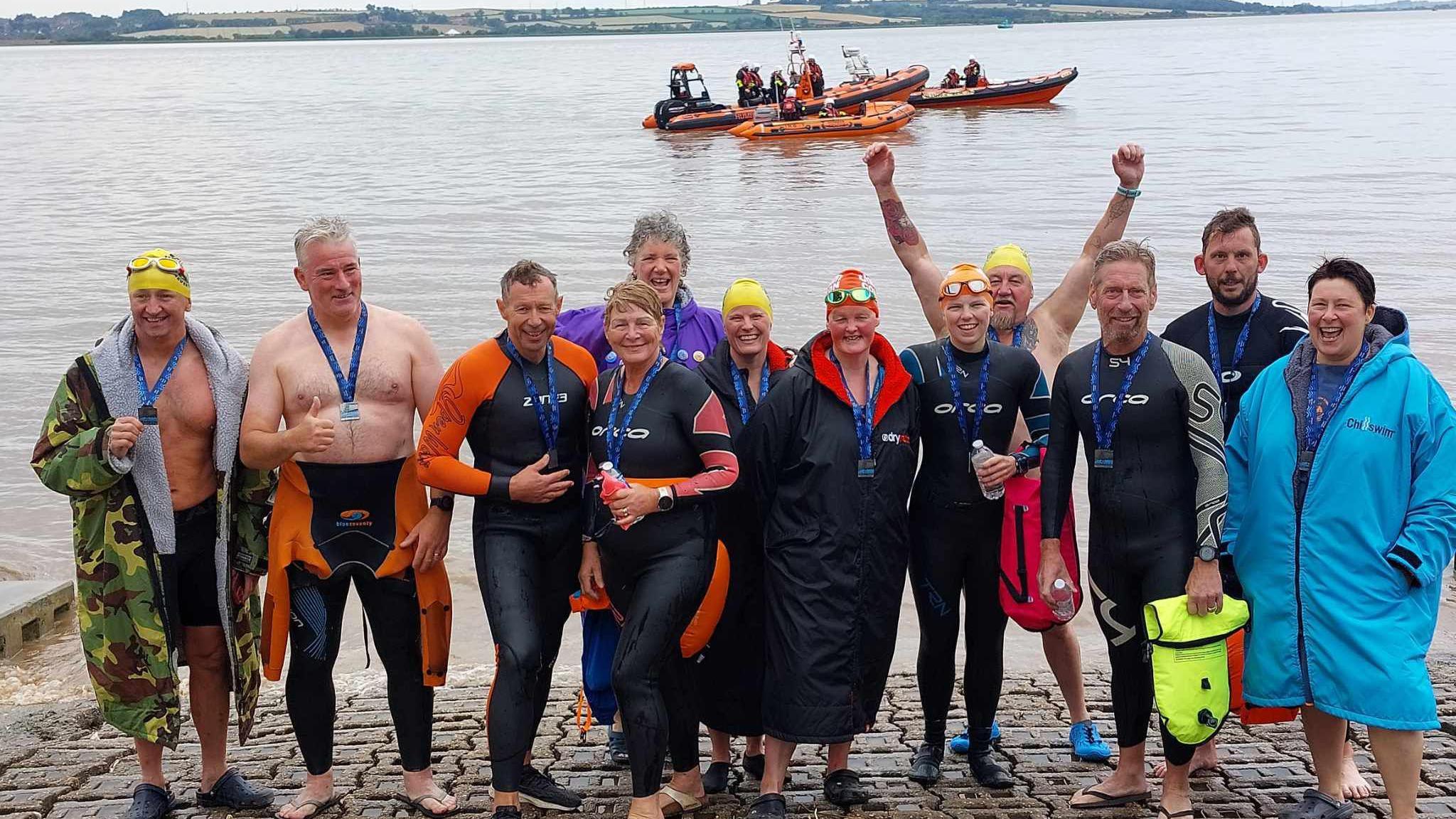 Twelve swimmers dressed in swimming gear standing on a path in front of the Humber. In the distance are three orange lifeboats with workers on board