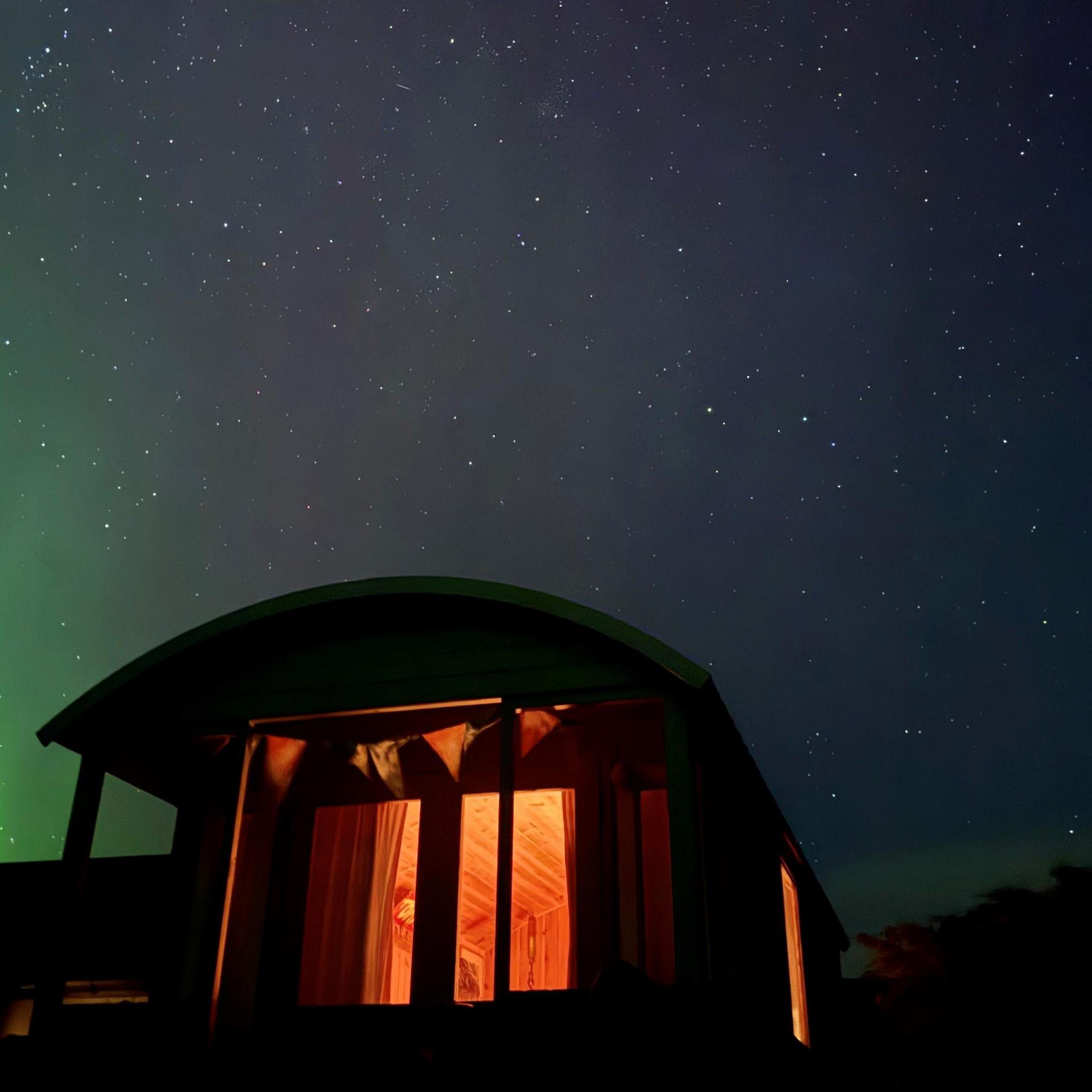The lights from a shepherds hut, which is decorated with bunting, provide a warm glow as the starry night sky above plays host to a colourful aurora.