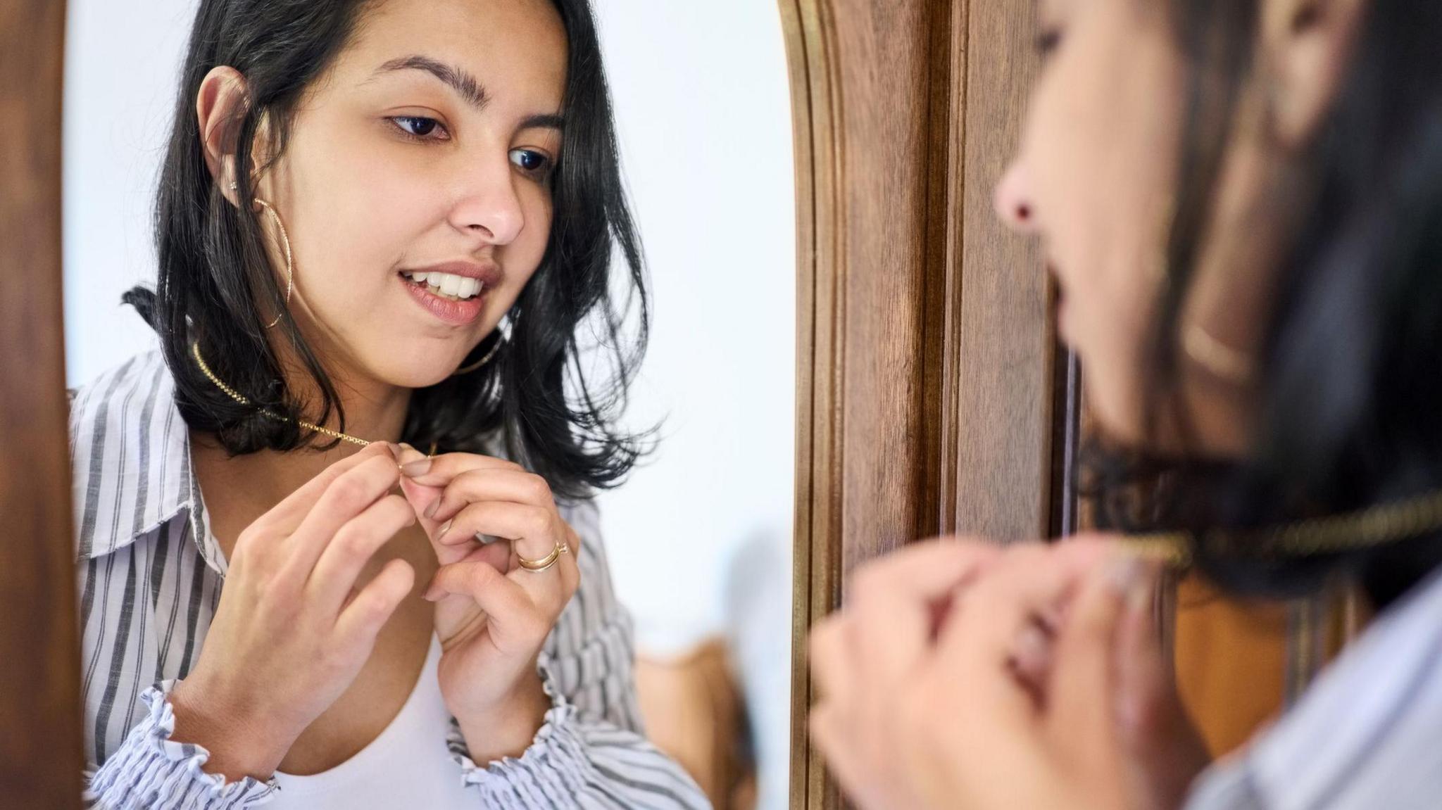 A woman puts on a gold necklace while looking in the mirror