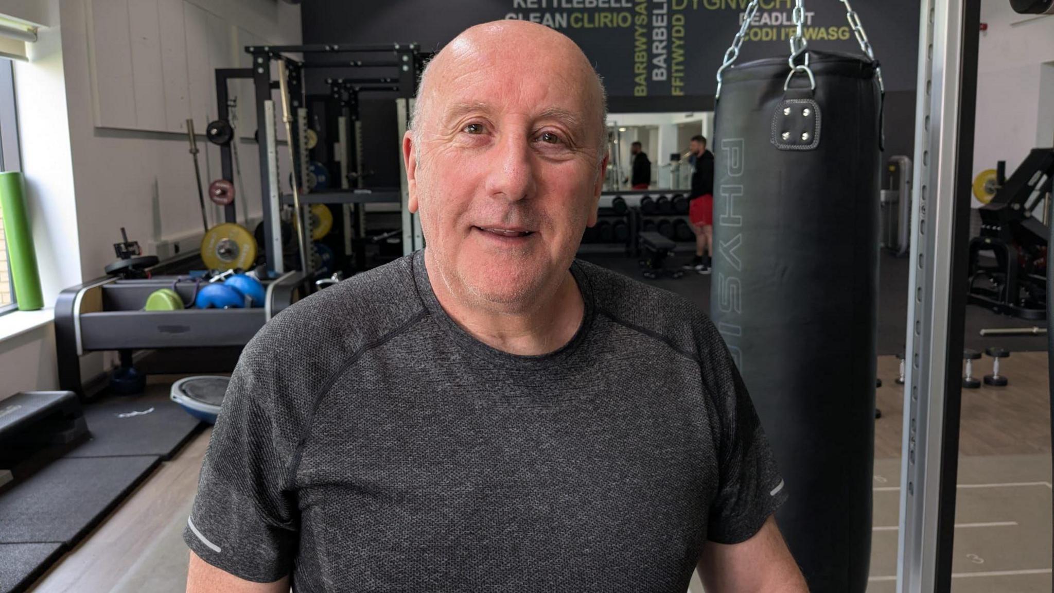 Paul is wearing a grey exercise top and is stood in a gym.