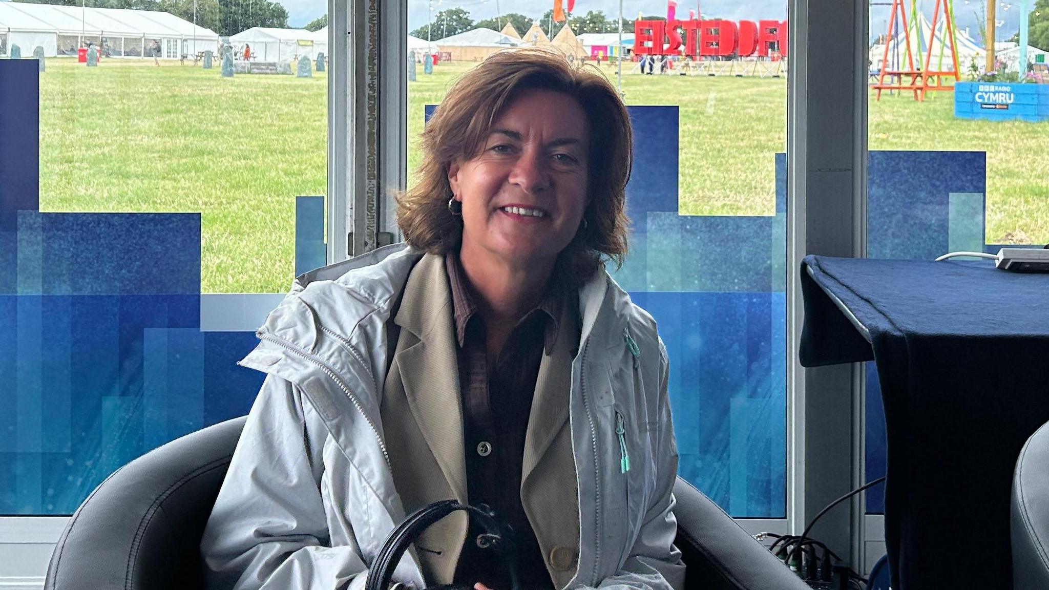 Eluned Morgan yn eistedd yn stiwdio Radio Cymru. Mae'n gwisgo cot law gwyn ac yn dal par o glustffonau.