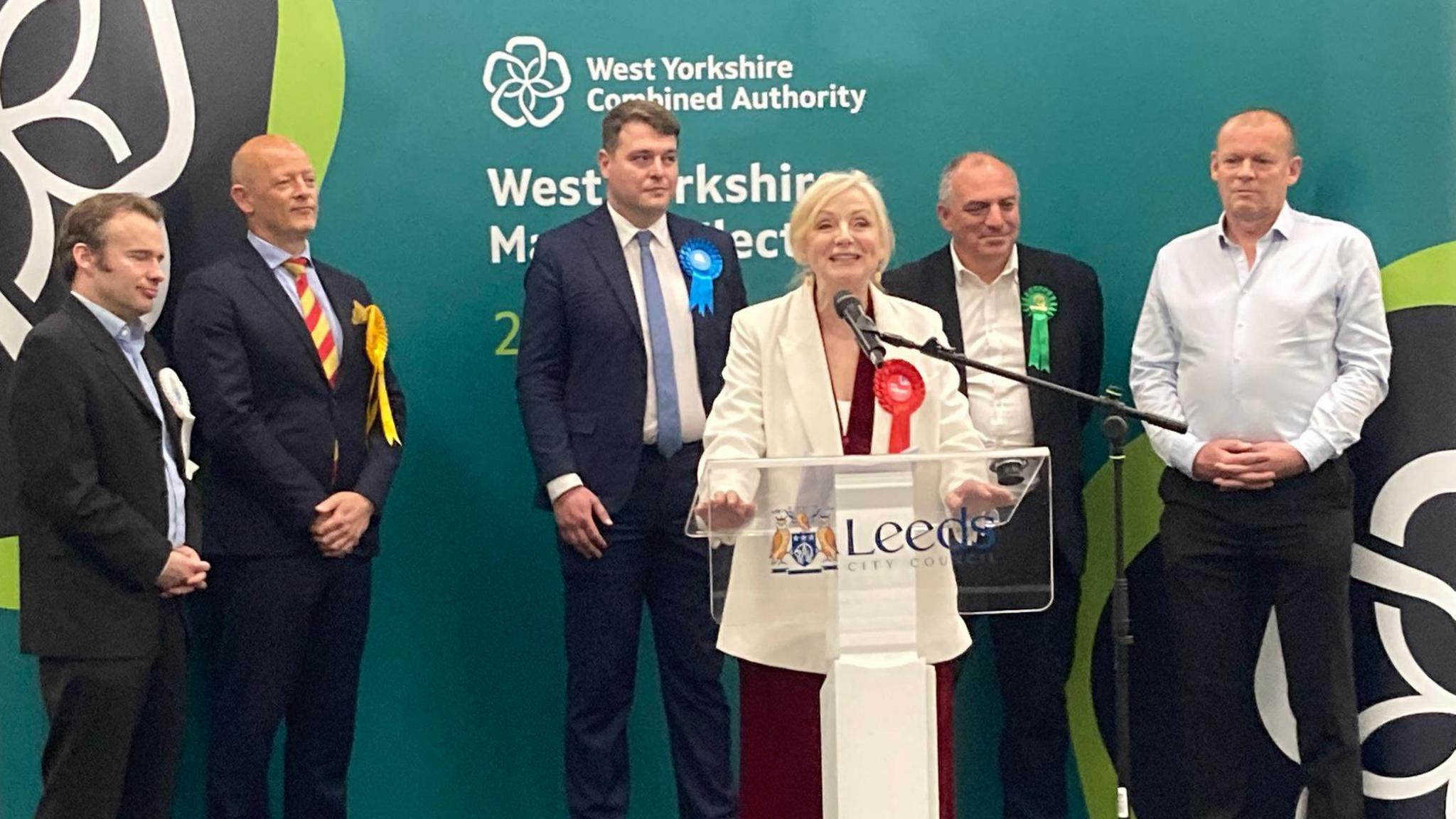 Mayor Brabin is stood at a podium making wearing a white jacket with a red Labour rosette on it. The other five candidates are in the background listening to her re-election speech. 