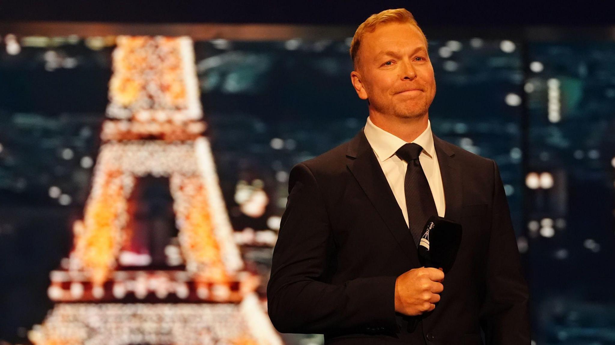 Sir Chris Hoy, during the 2024 BBC Sports Personality of the Year Awards held at MediaCityUK, Salford, wearing a black suit and tie with white shirt and holding a microphone. Behind him is an out of focus Eiffel Tower.